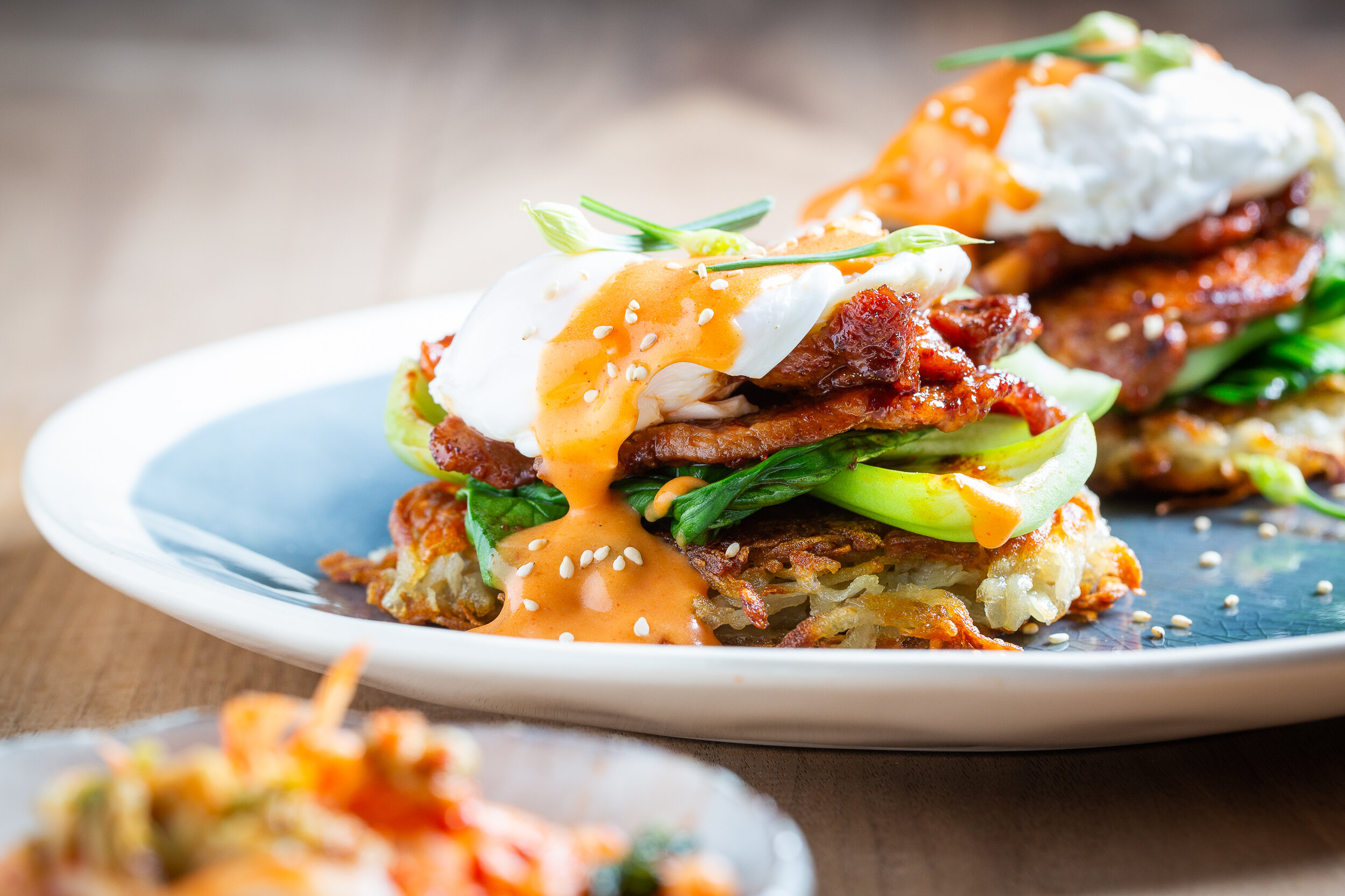 Breakfast dish with poached eggs, bacon, avocado on hash browns, drizzled with sauce and sesame seeds, on a plate.