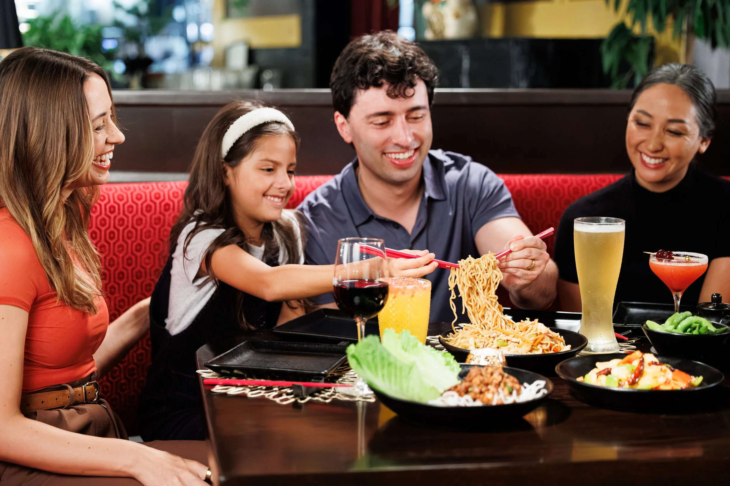 Huge Galdones food and lifestyle photography for P.F. Chang's showing a multi-generational family of three adults and a child, sitting around a table at a restaurant with various Asian dishes and drinks, smiling and enjoying their meal.