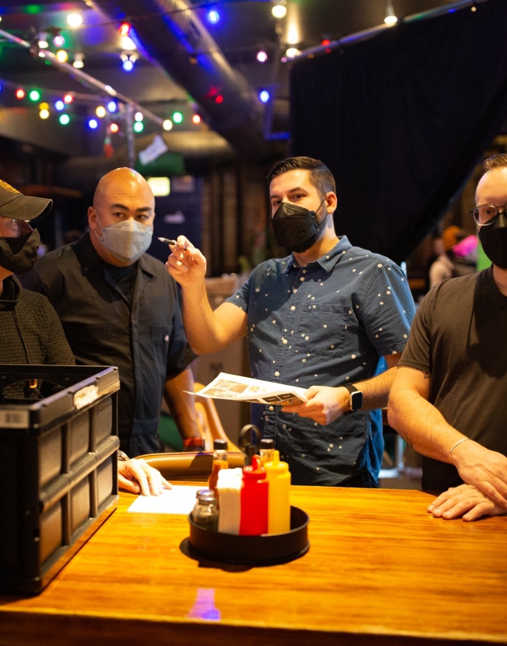 Group of men wearing face masks in a restaurant, holding papers and looking engaged in conversation, with colorful string lights in the background.