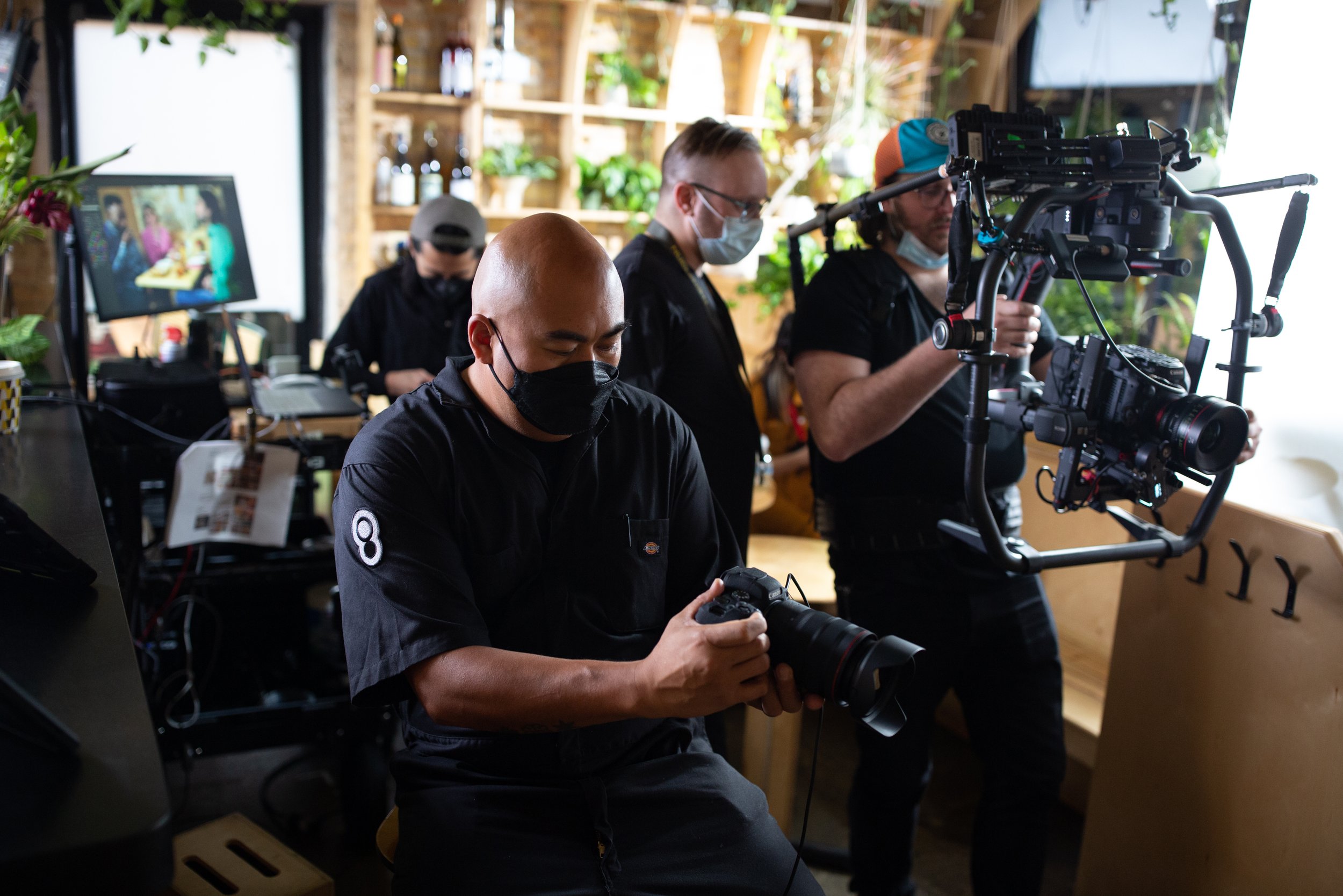 Film crew working with cameras and equipment in a studio setting, wearing masks.