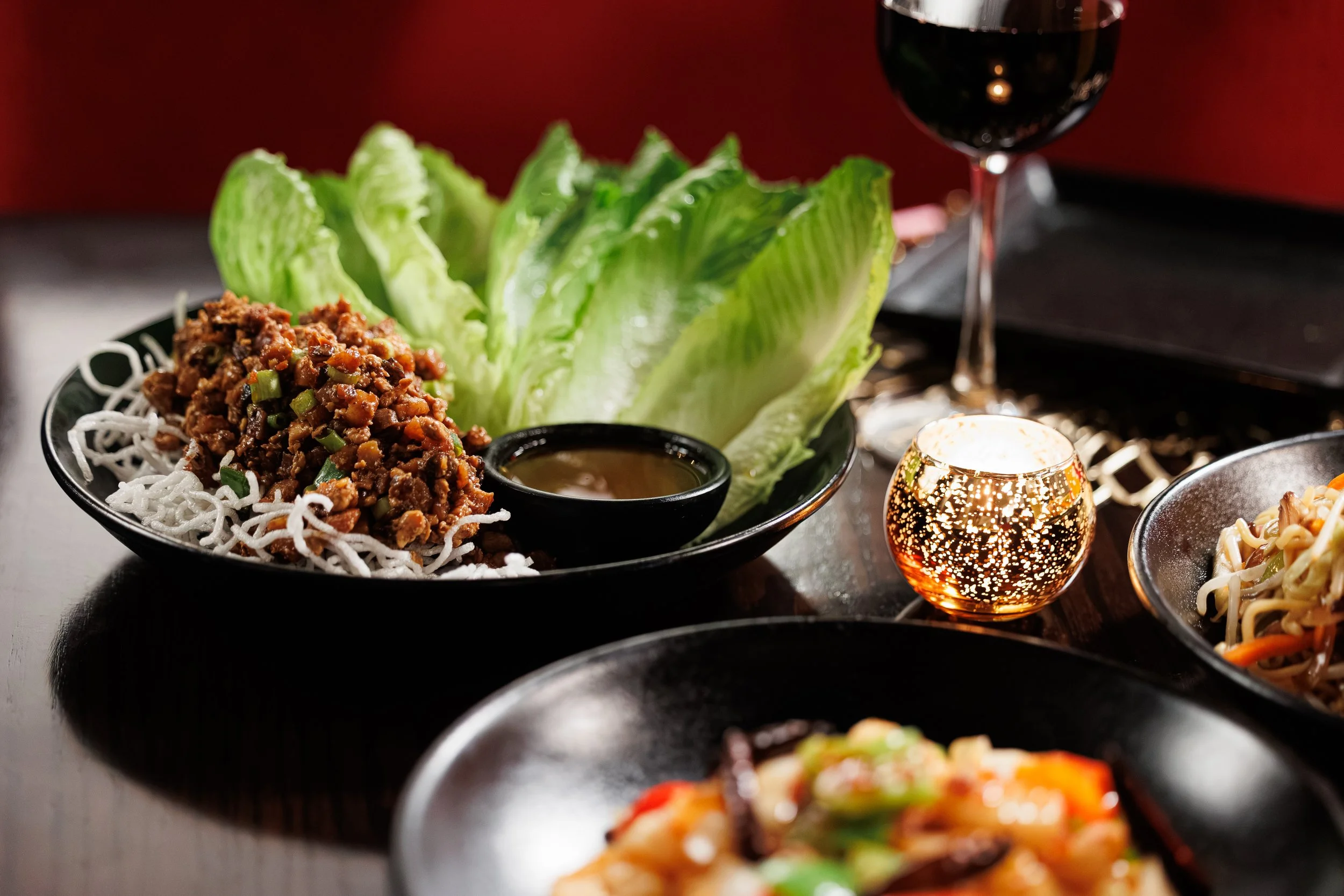 Huge Galdones  lifestyle,  foodservice and restaurant photography for P.F. Chang's showing a plate of Asian food featuring rice noodles, a chili, and a seasoned ground meat dish with lettuce, with a glass of red wine in the background.