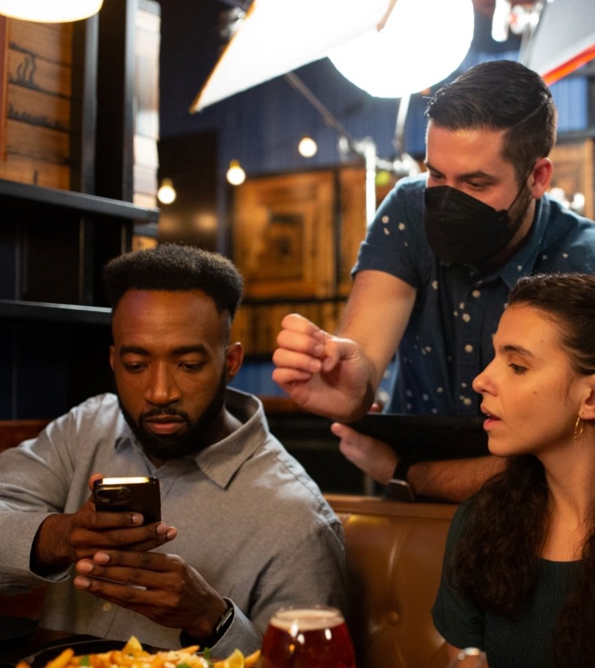 Group of people in a restaurant setting, one person using a smartphone, another pointing at the phone. Food and drink are on the table.