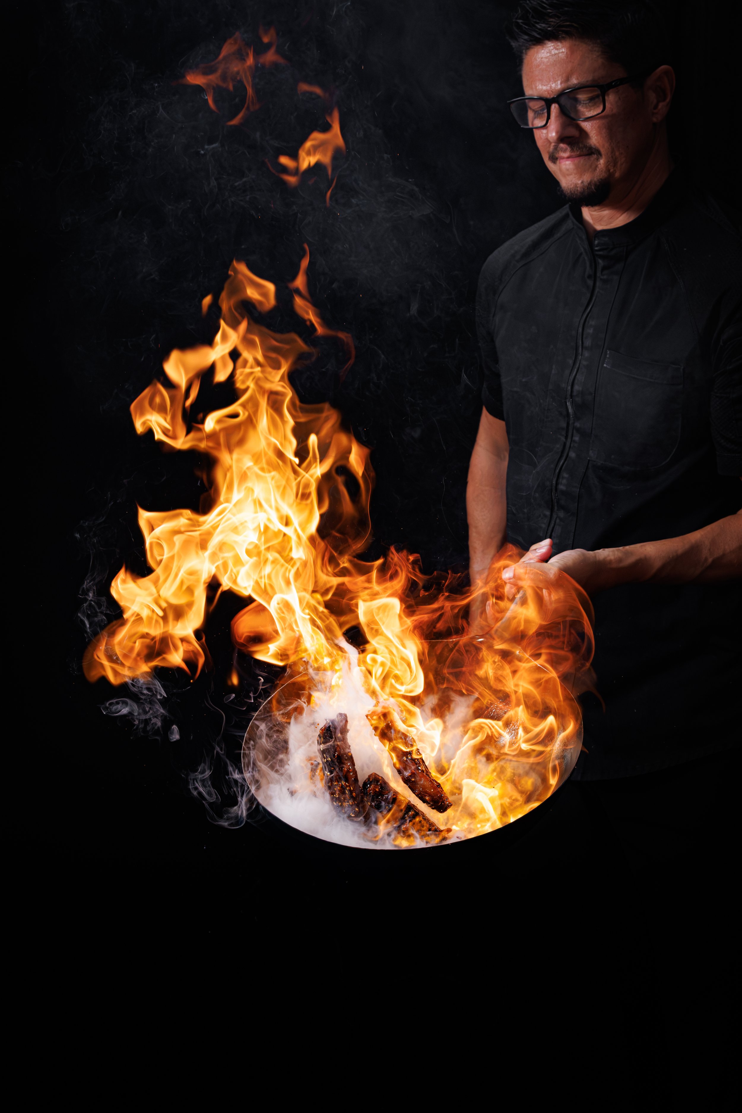Huge Galdones food and lifestyle photography for P.F. Chang's of a Chef in a black jacket shirt flipping ribs in a wok over a large fire on a dark background.