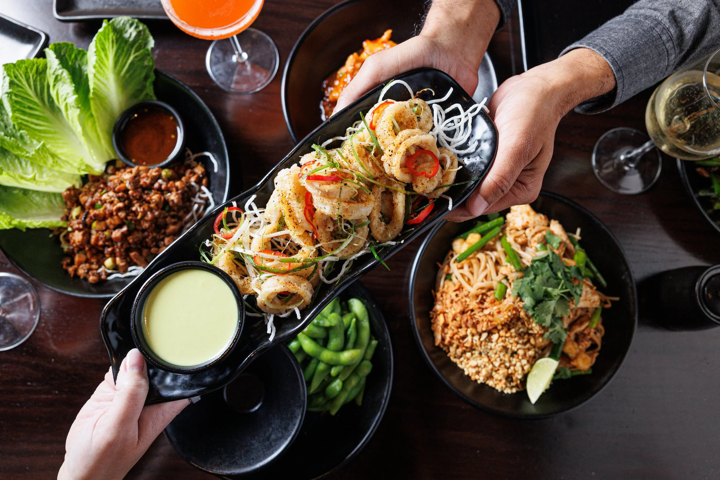Huge Galdones  lifestyle, foodservice and restaurant photography for P.F. Chang's showing a hand holding a black oval plate with fried calamari garnished with sliced red chili and green herbs, surrounded by other Asian dishes.