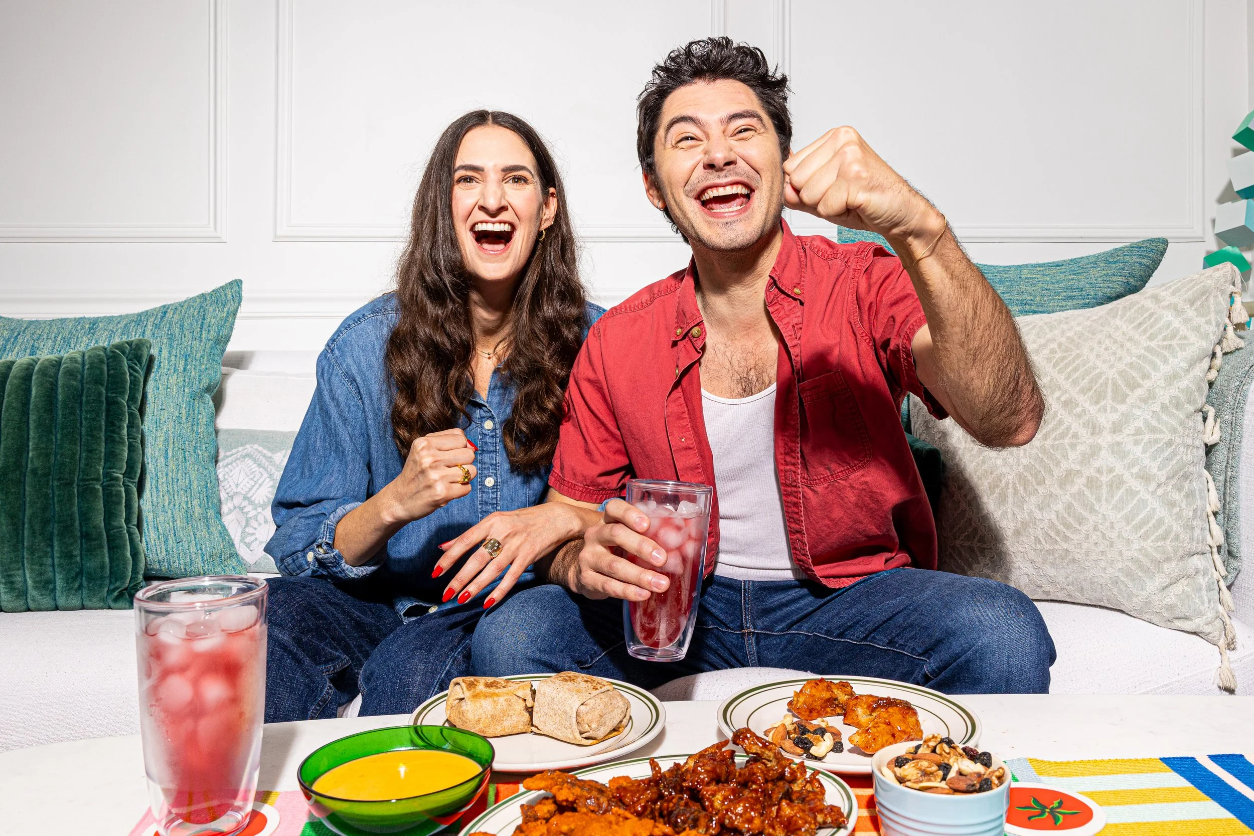A happy couple celebrating with food and drinks on a white couch, surrounded by colorful pillows.