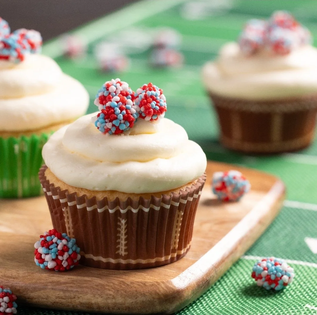 Huge Galdones photograph for Nerds Gummy Clusters cupcake with white frosting topped with red, white, and blue sprinkles, placed on a wooden board with scattered sprinkles around.