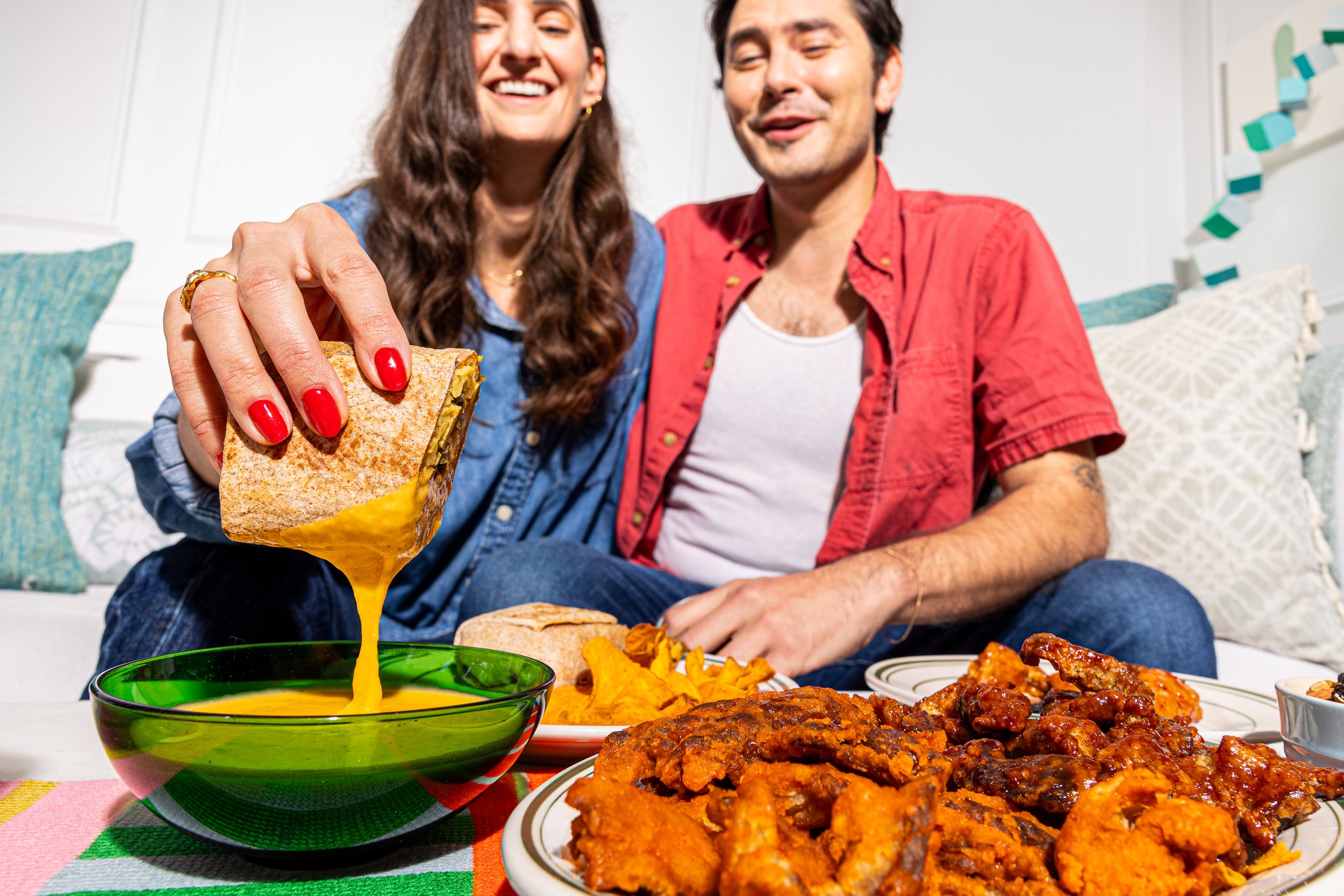 A woman with red nail polish and a man are sitting at a table with Mexican food, including a bowl of cheese dip, vegan fried chicken, and nachos. The woman is holding a burrito with cheese, and both are smiling.