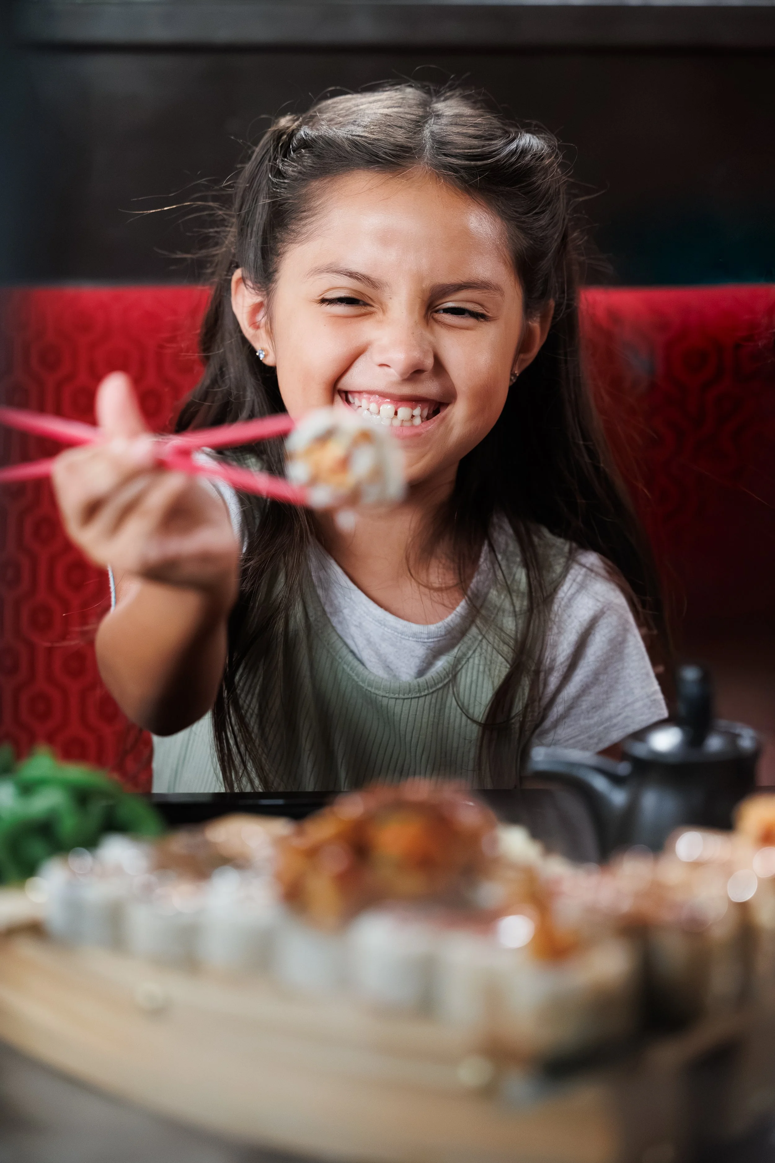 Huge Galdones food and lifestyle photography for P.F. Chang's showing a young girl with dark hair smiling while holding a piece of sushi with red chopsticks.