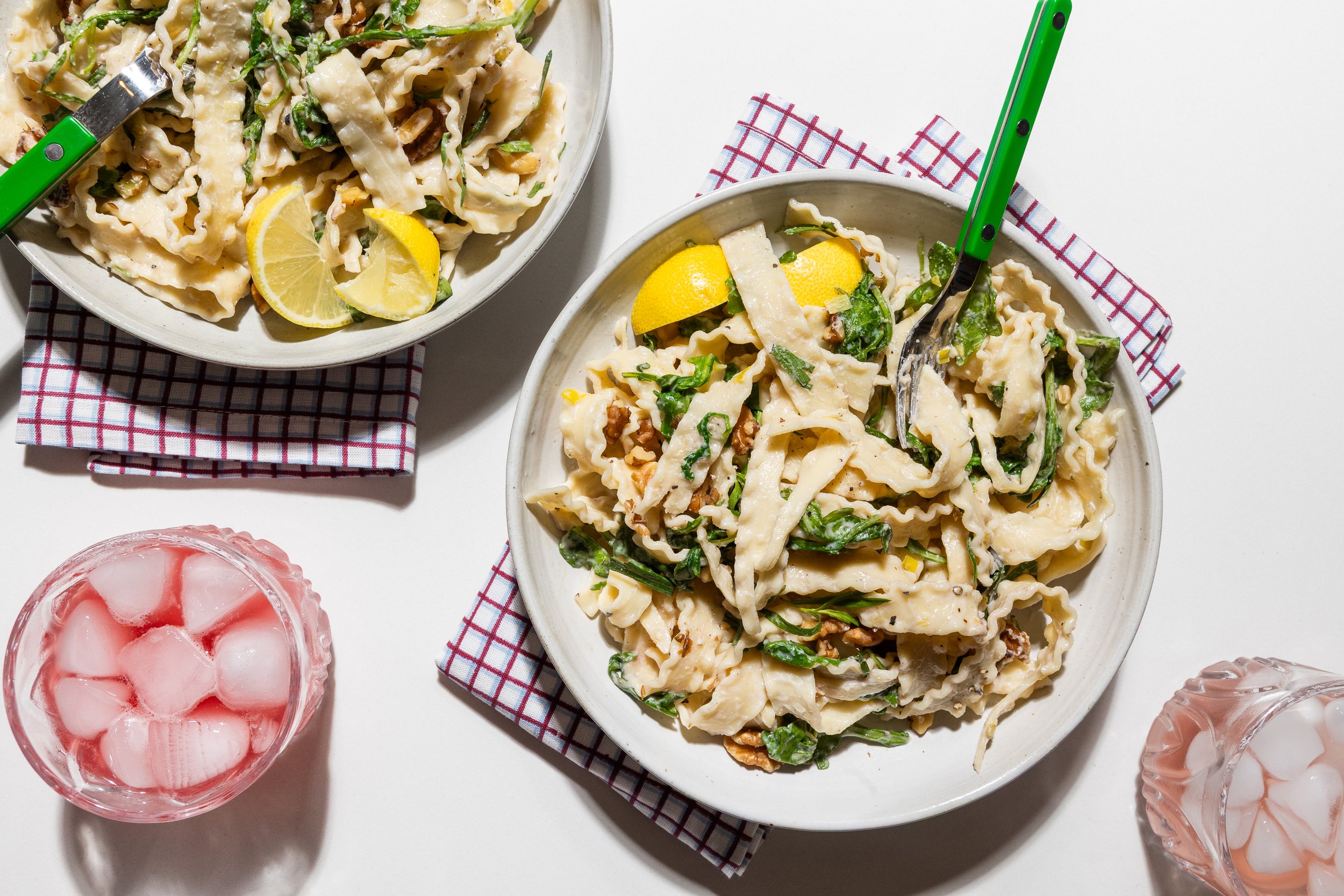 Two bowls of pasta salad, with lemon wedges, on checkered napkins, with two glasses of pink iced drinks on a white surface.