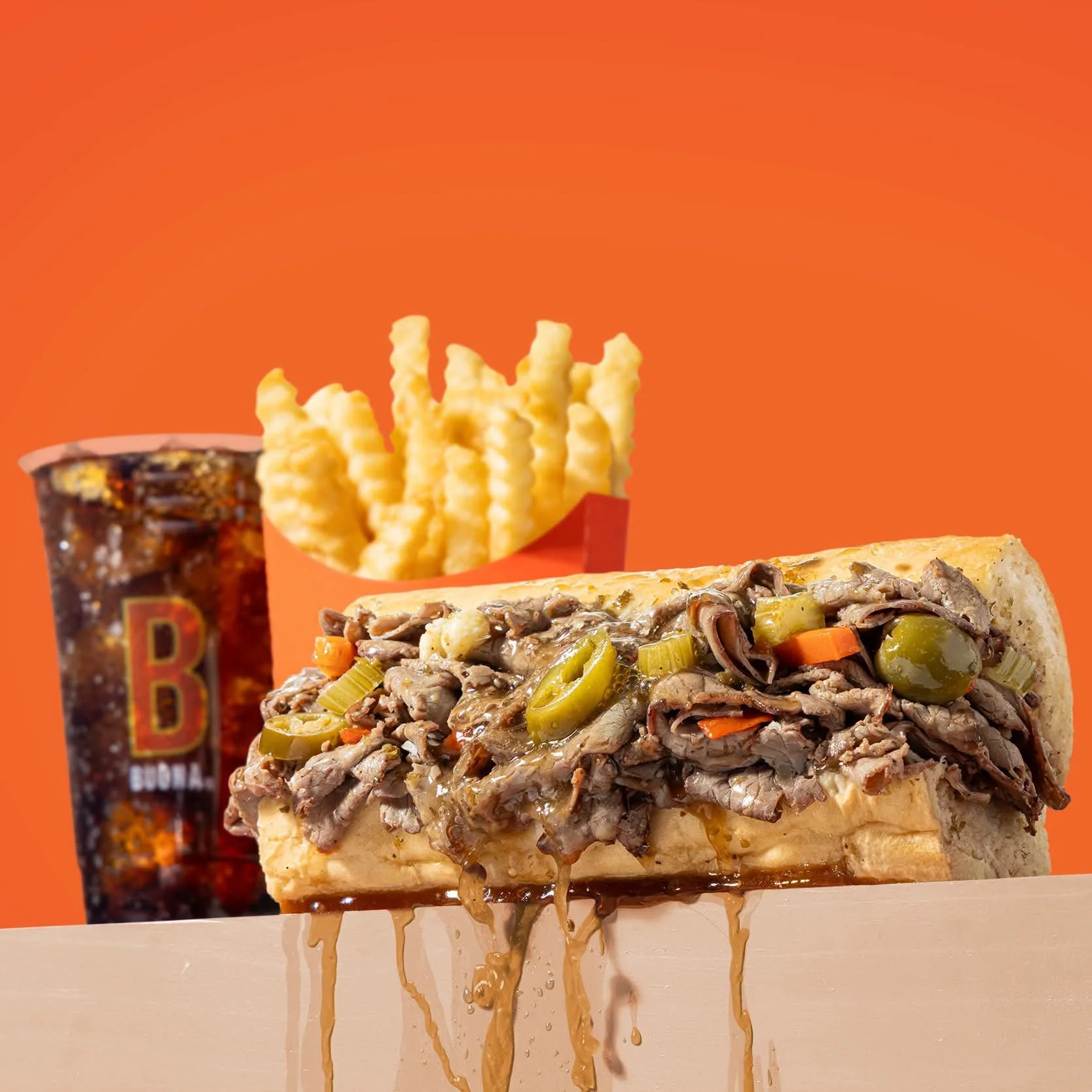 A cheesesteak sandwich with sliced beef, green peppers, and onions, topped with melted cheese, dripping juices, and sauce on a wooden surface. In the background, there are crinkle-cut French fries in a red container, a soda with a colorful label, and a slice of pizza with mushroom toppings, all against an orange background.