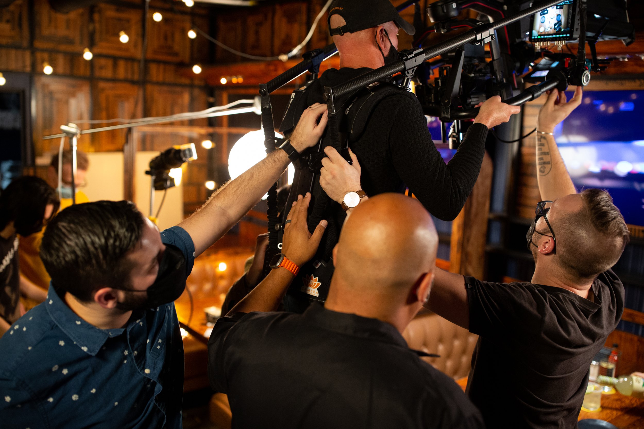 Film crew setting up camera equipment, adjusting a camera rig with bright lighting in the background, in a studio setting.