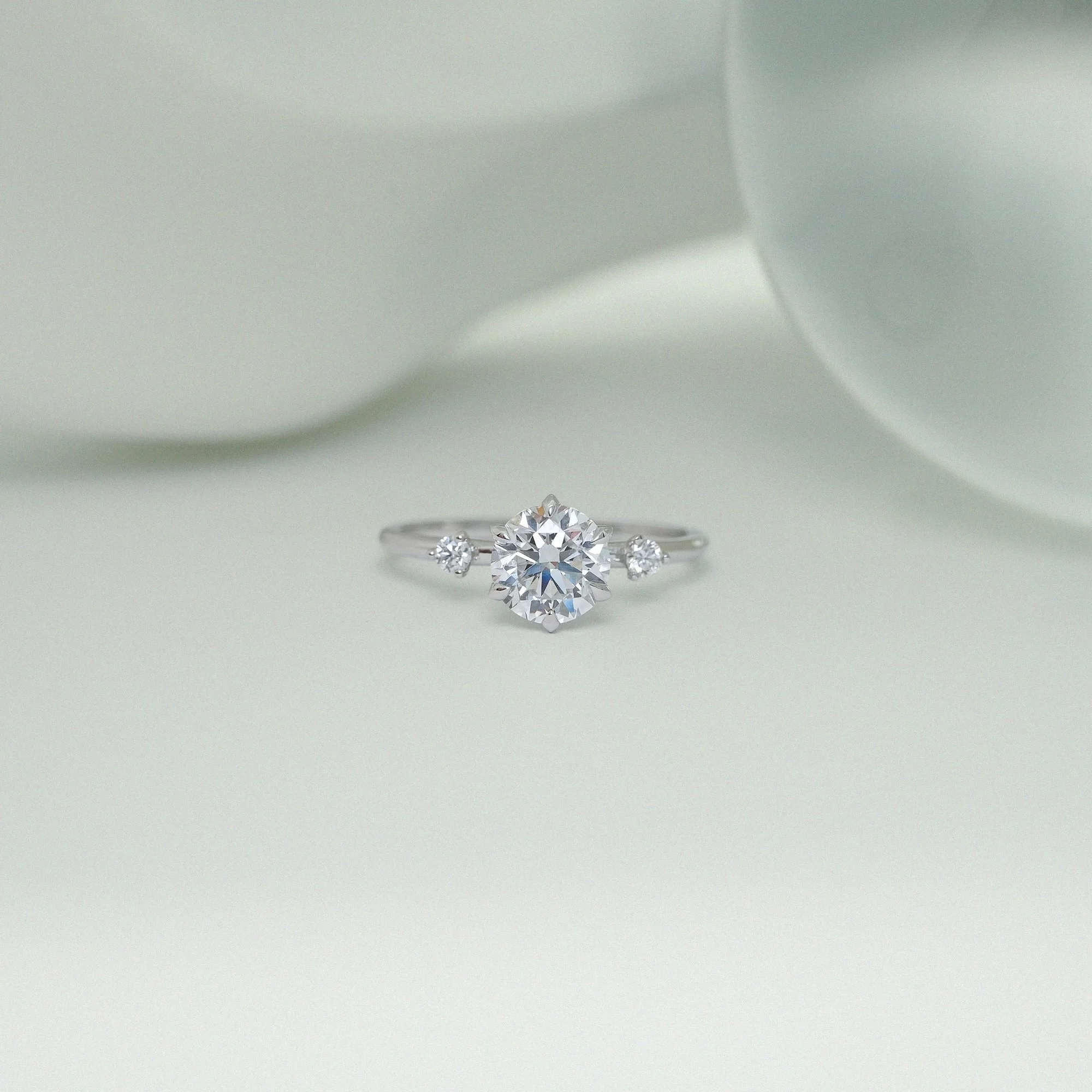 A diamond that blooms like a flower - The Amelia Ring centres on a round brilliant-cut diamond, framed by two delicately spaced diamonds along the shoulders, creating a sense of balance, light, and gentle rhythm. A graceful six-claw lotus-inspired se