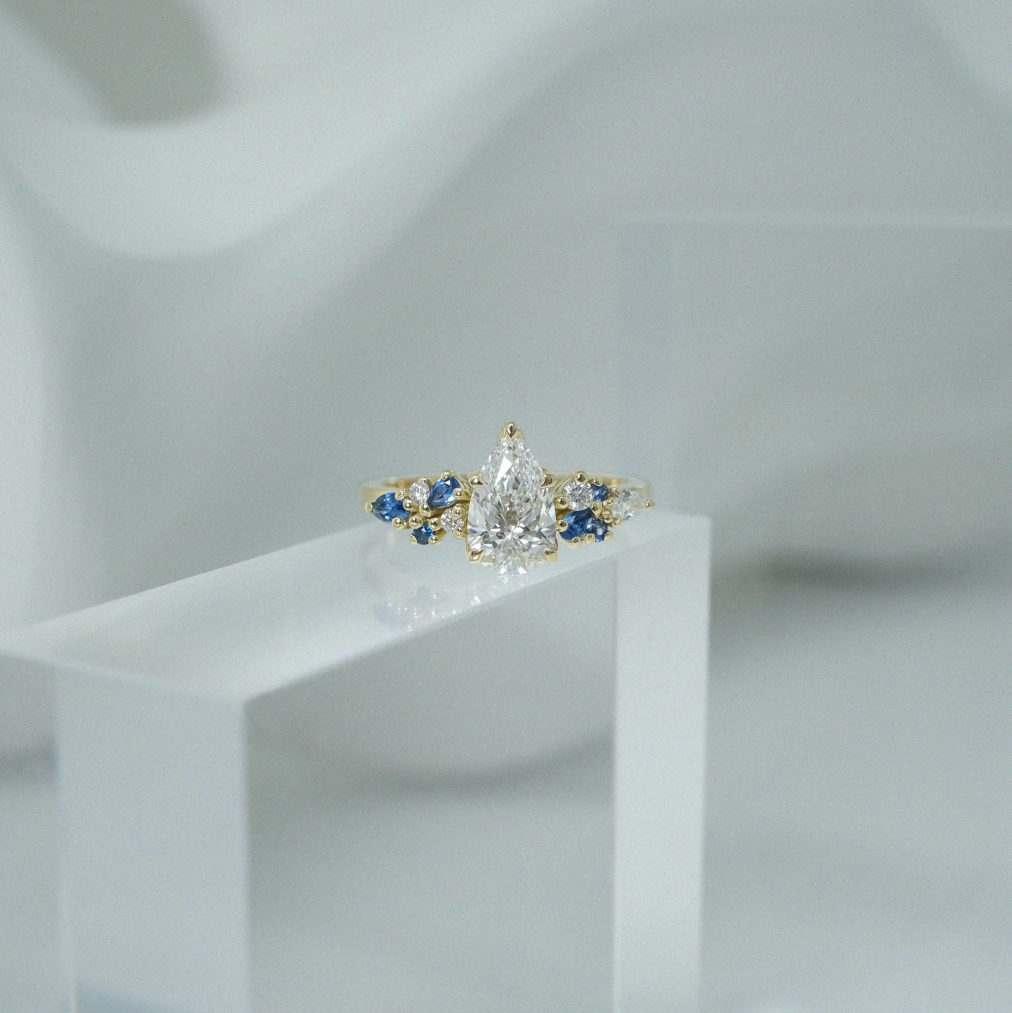 Untamed and beautifully off balance. The Ilias Ring features a pear-cut diamond held in a delicate five-claw floral-inspired setting, its soft curves echoing the elegance of blooming petals. Along the shoulders, pear and round brilliant-cut sapphires