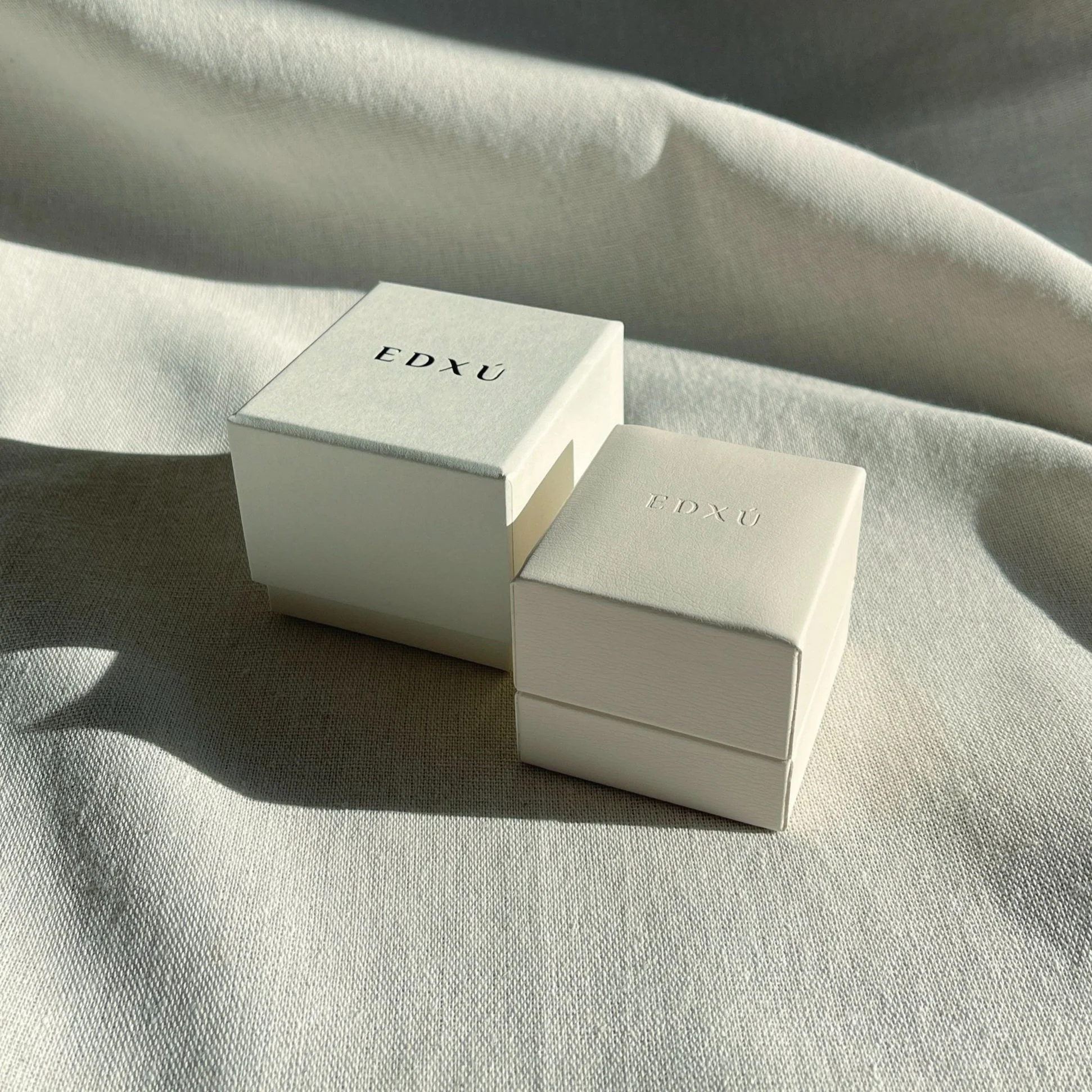 Two white jewelry boxes with the brand name 'EDXŪ' on a light fabric surface, lit by sunlight, casting shadows.