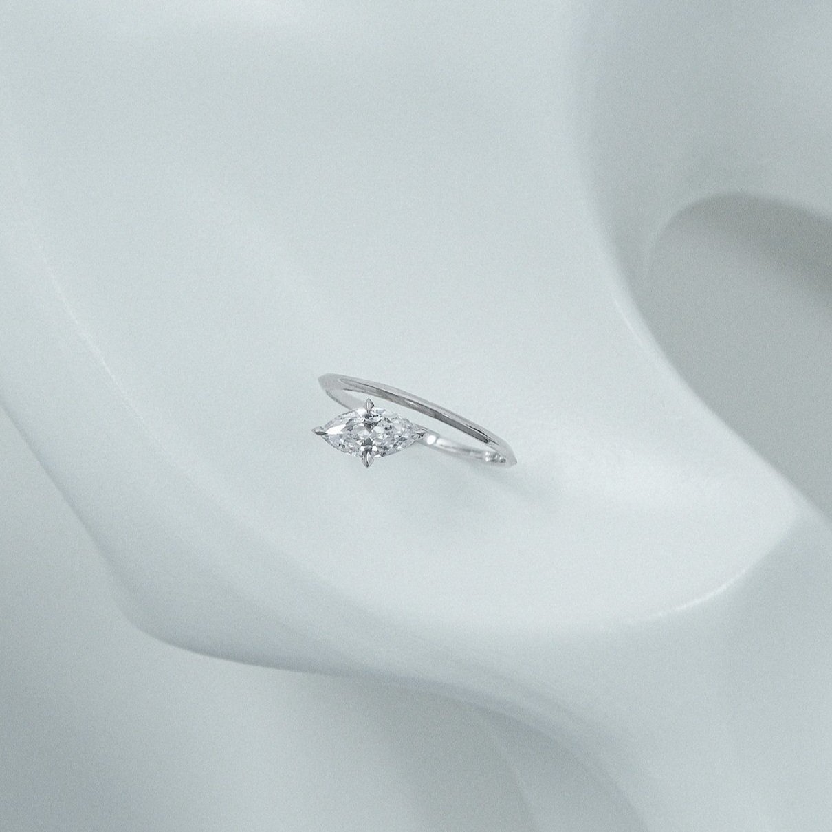 An asymmetrical platinum engagement ring with a marquise-cut diamond on a light, curved surface.