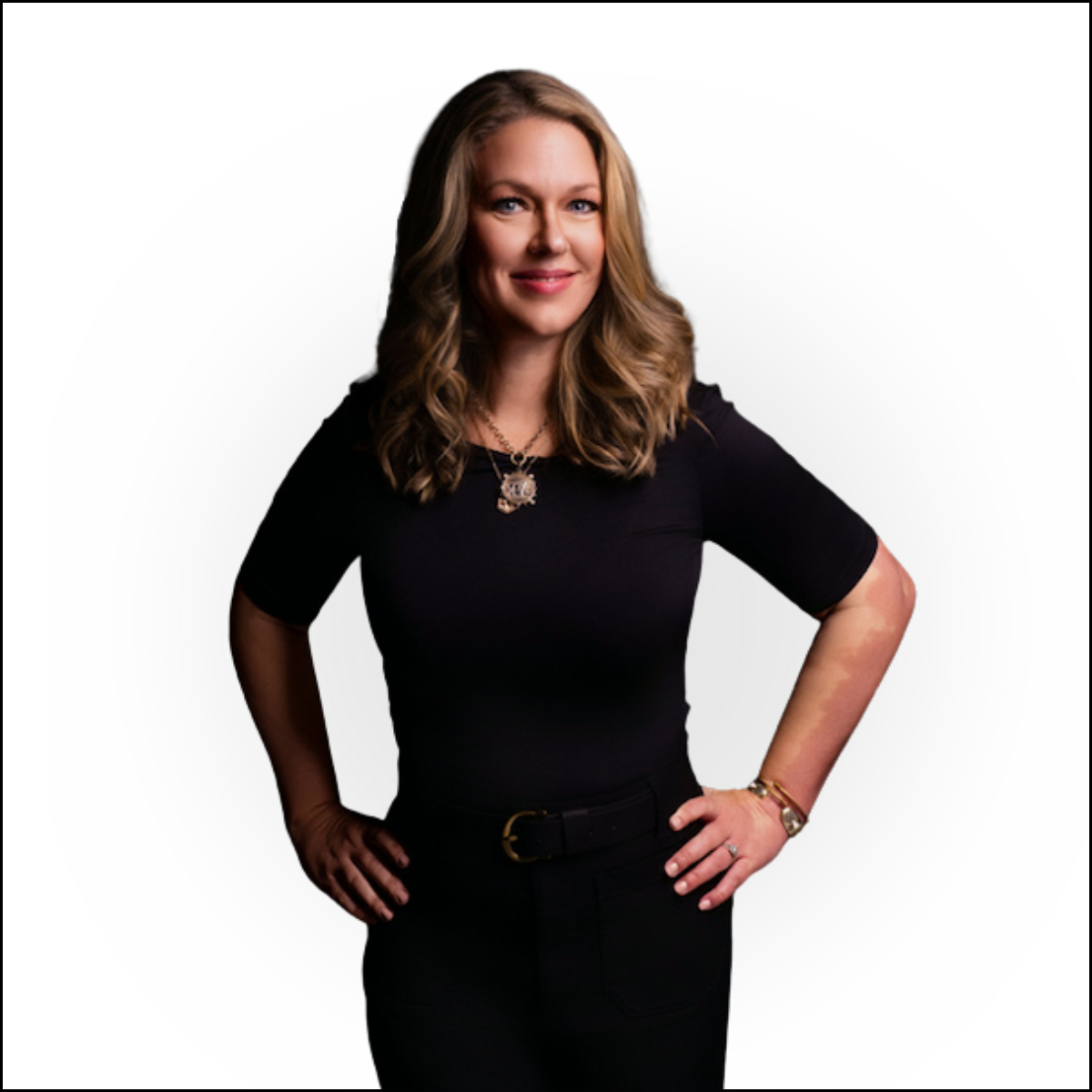 A woman with long wavy brown hair wearing a black dress with short sleeves, accessorized with a necklace, bracelet, and watch, standing with hands on her hips against a white background.