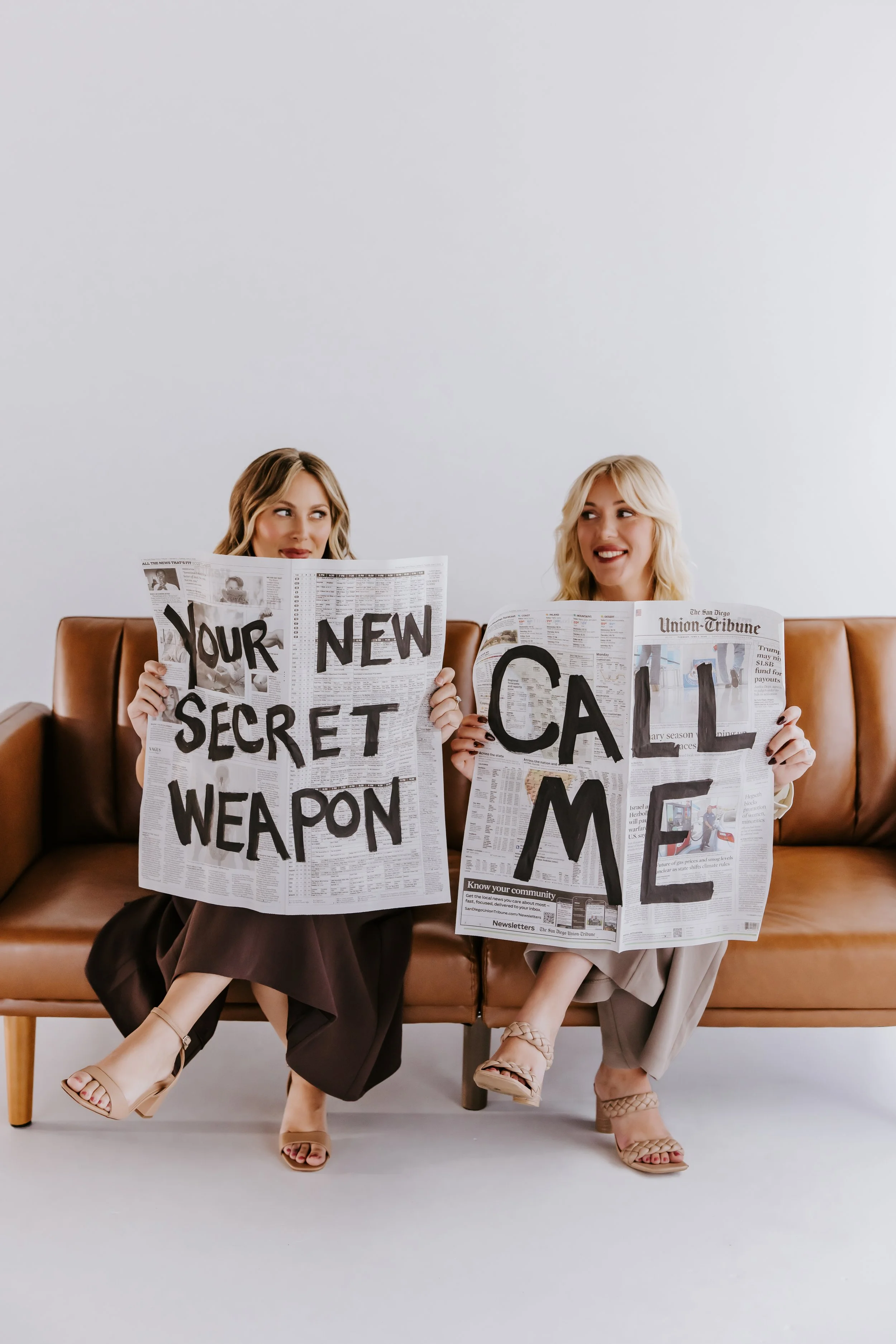 Two women sitting on a brown leather couch, each holding a newspaper with large, bold words on the front pages. The woman on the left has light brown hair and is wearing a brown skirt and beige high-heeled sandals, holding a newspaper that reads "YOUR NEW SECRET WEAPON." The woman on the right has blonde hair and is wearing beige pants and beige sandals, holding a newspaper that reads "CALL" with additional text partially visible.