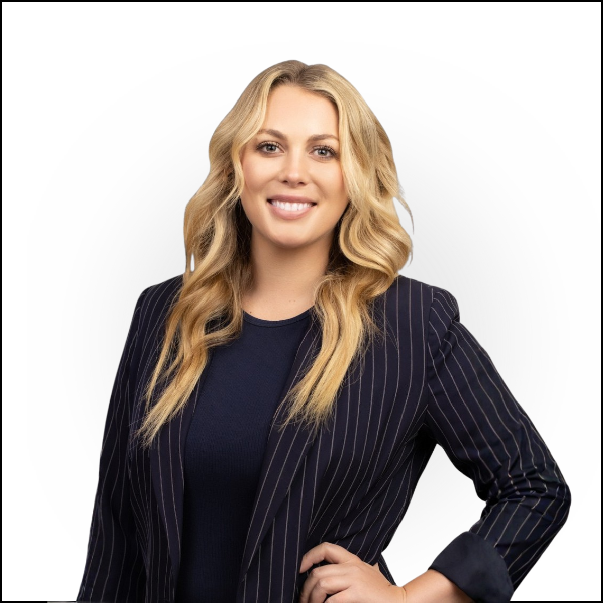A woman with long blonde hair in loose waves, wearing a navy pinstripe blazer over a navy top, smiling and standing with one hand on her hip against a white background.