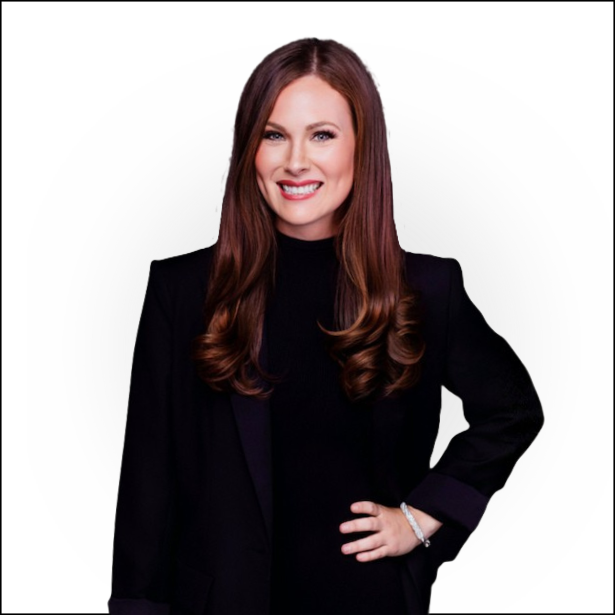 Portrait of a woman with long wavy red hair, wearing a black blazer and black top, smiling with one hand on her waist against a white background.