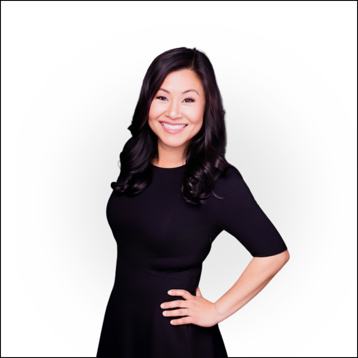 A woman with long black hair smiling and posing with her hand on her hip, wearing a black dress, against a white background.