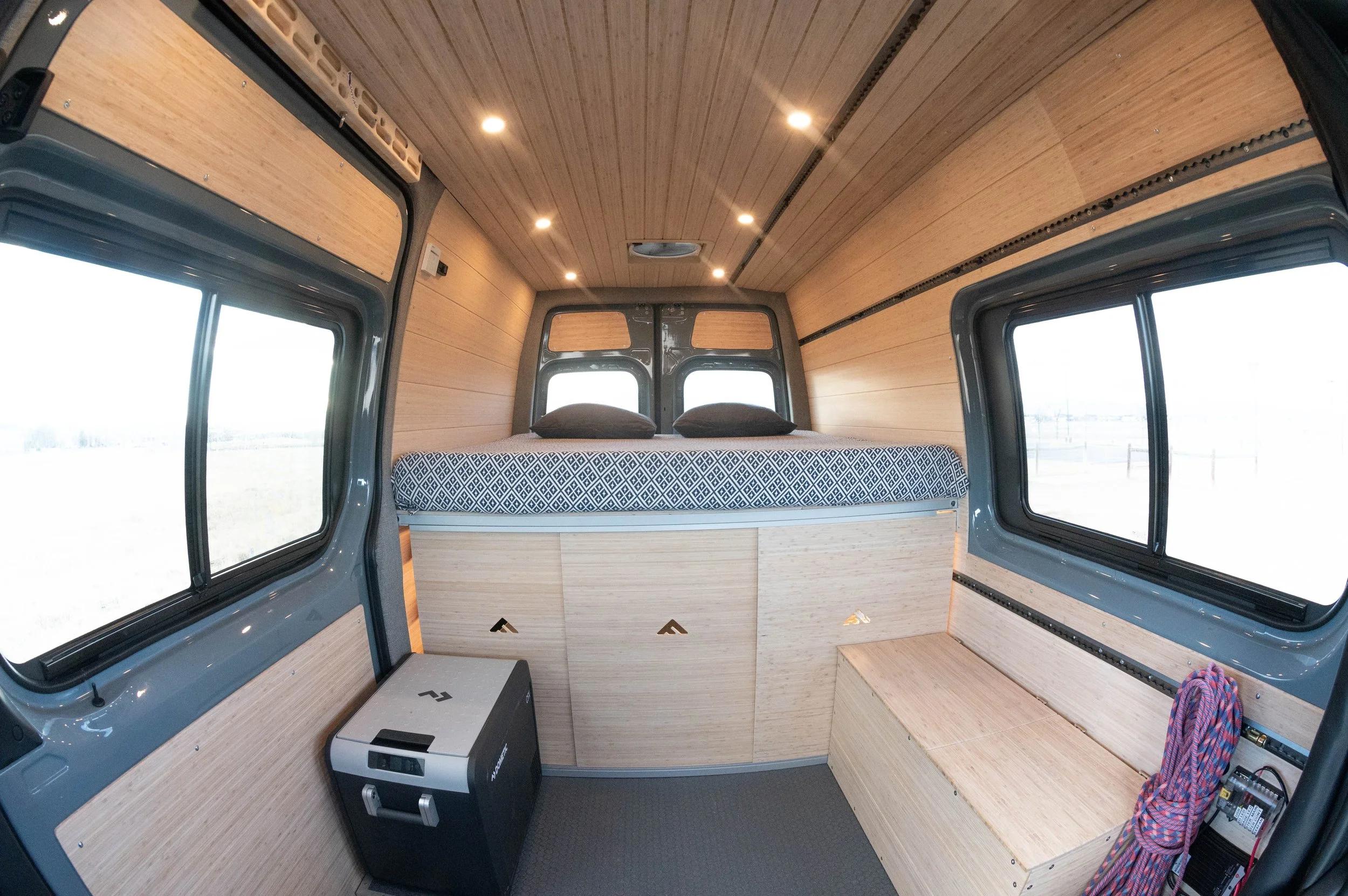 Interior of a camper van with wooden paneling, a bed with black pillows, windows on three sides, a small refrigerator, and a bench with a rope on the right side.