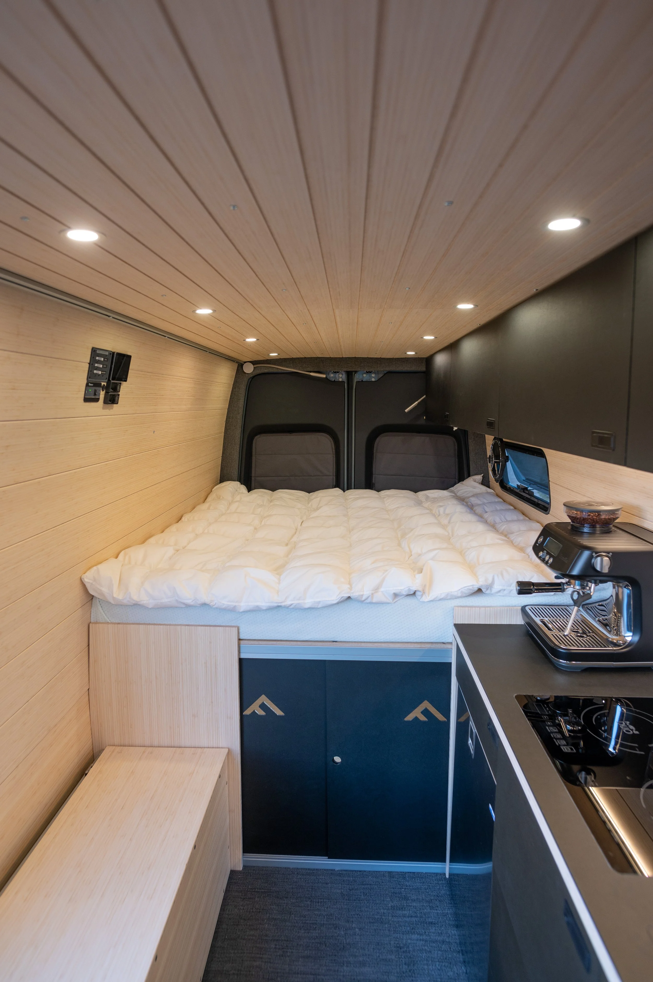 Interior of a small camper van with wood paneling, a bed with a white comforter, a coffee maker, and a small kitchen area.