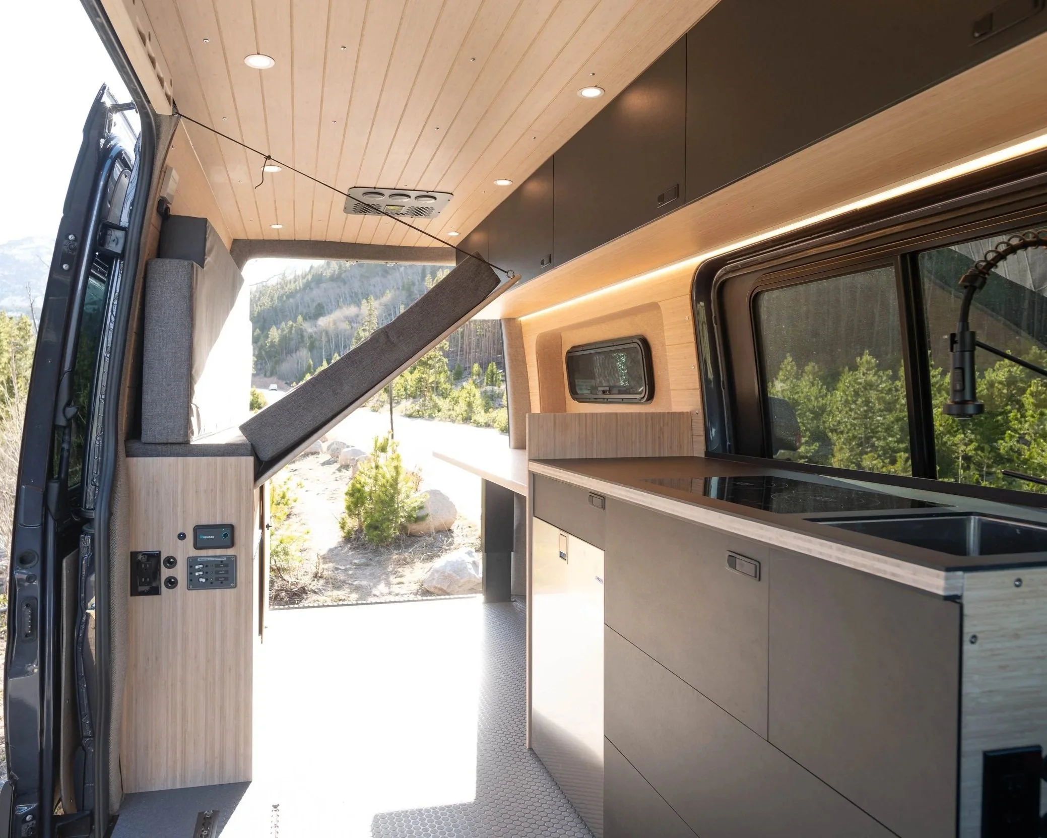 Interior of a modern camper van with wood-paneled ceiling and walls, black cabinets, and a window showing outdoor trees and mountains.