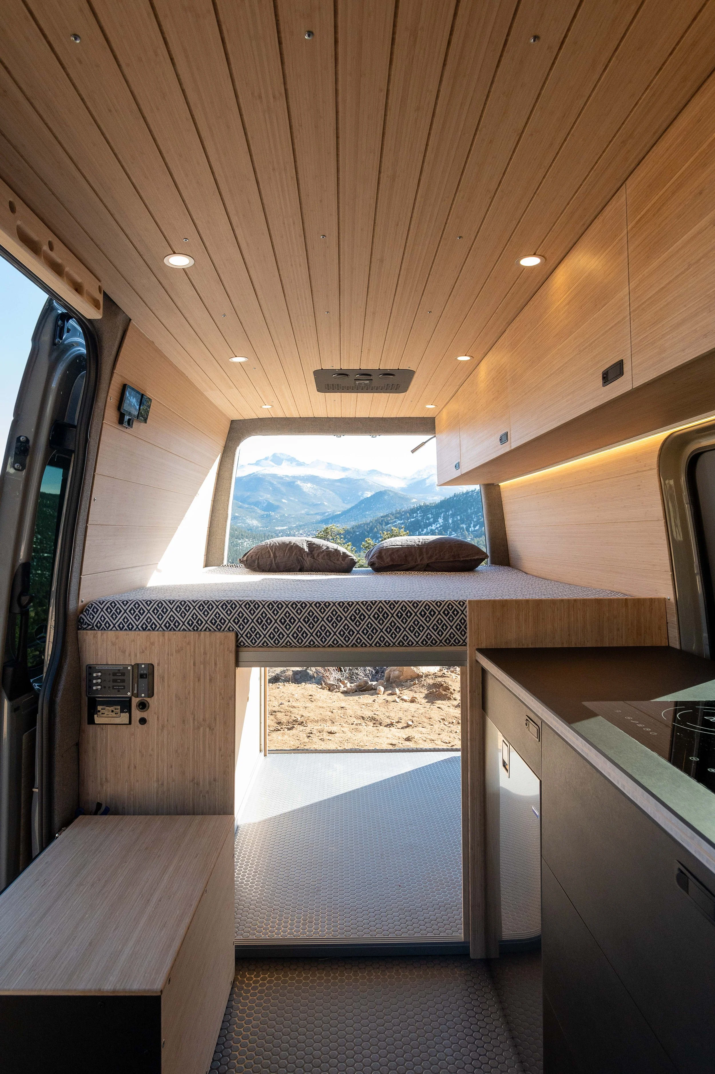 Interior of a converted camper van with a bed, wooden ceiling and walls, and a window showing mountain scenery outside.