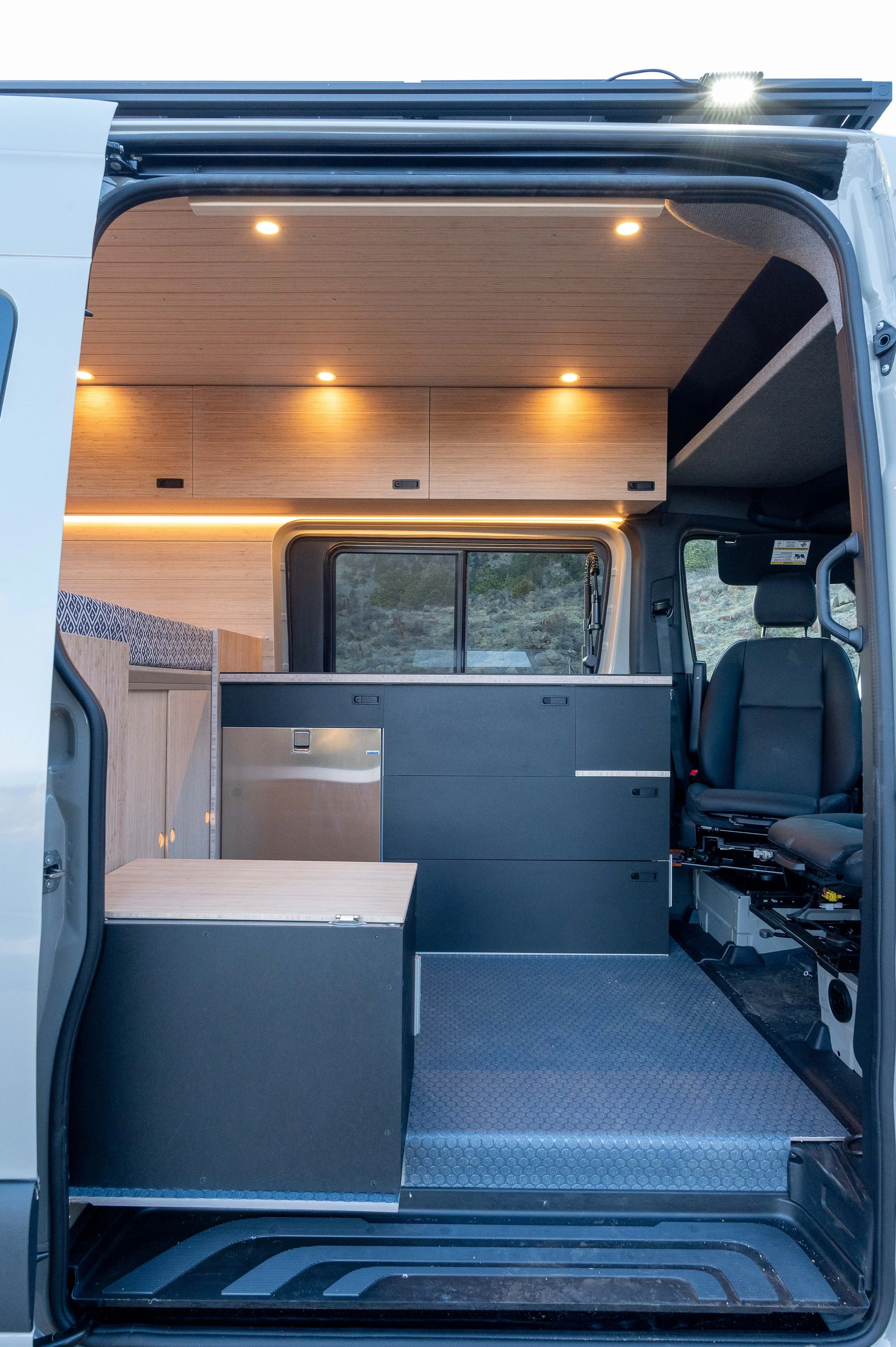 Interior of a camper van with wooden cabinetry, a seating area, and a driver's seat visible on the right.