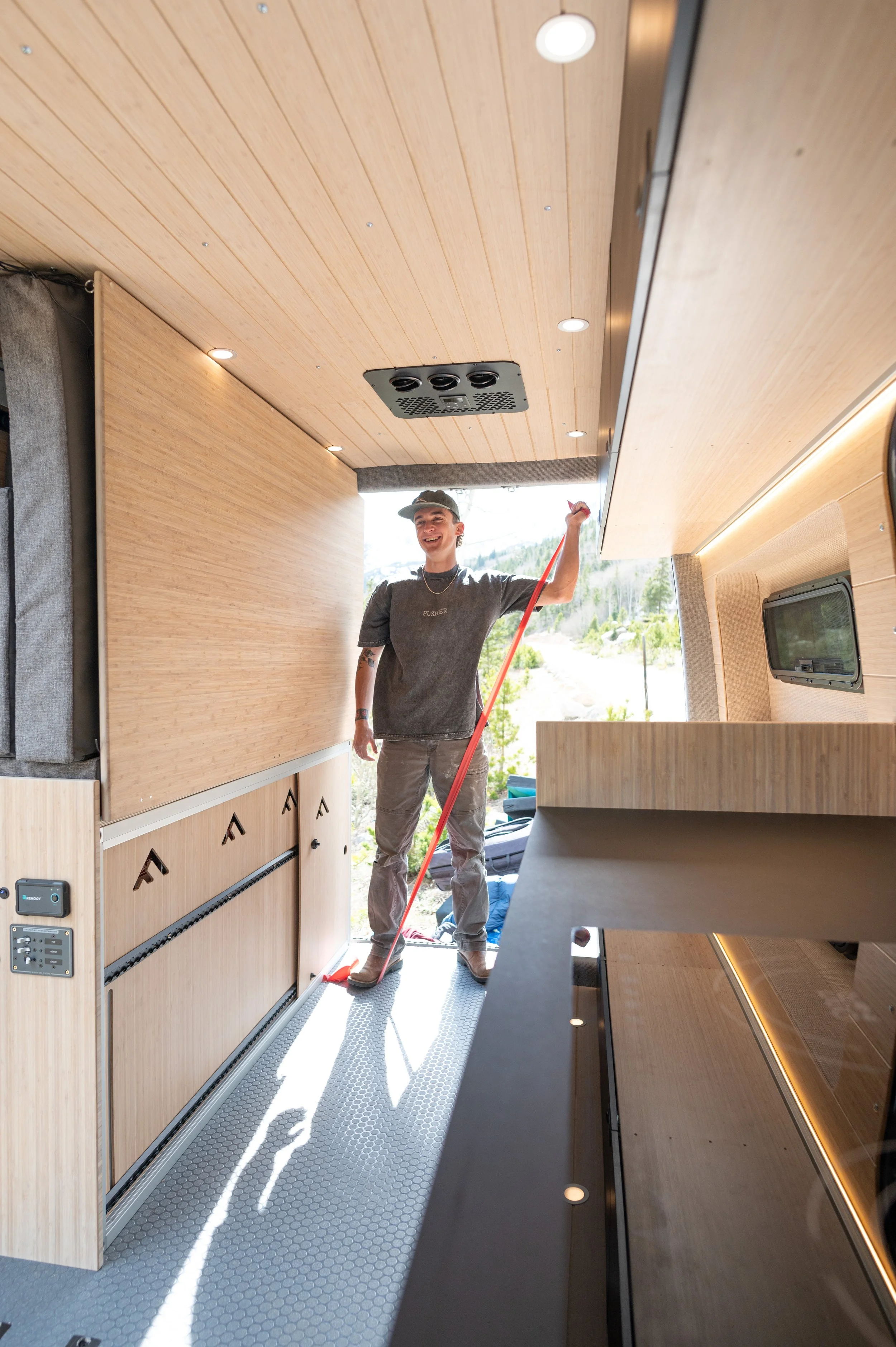 A person standing inside a wooden-paneled van or camper, mopping the floor, with a view of outdoor greenery and mountains through the back door.