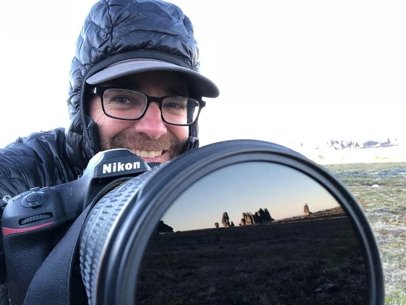 A man sitting at a table inside a tiny house or camper van, working on a laptop, with a DSLR camera and a water bottle beside him, and a landscape visible through the windows.