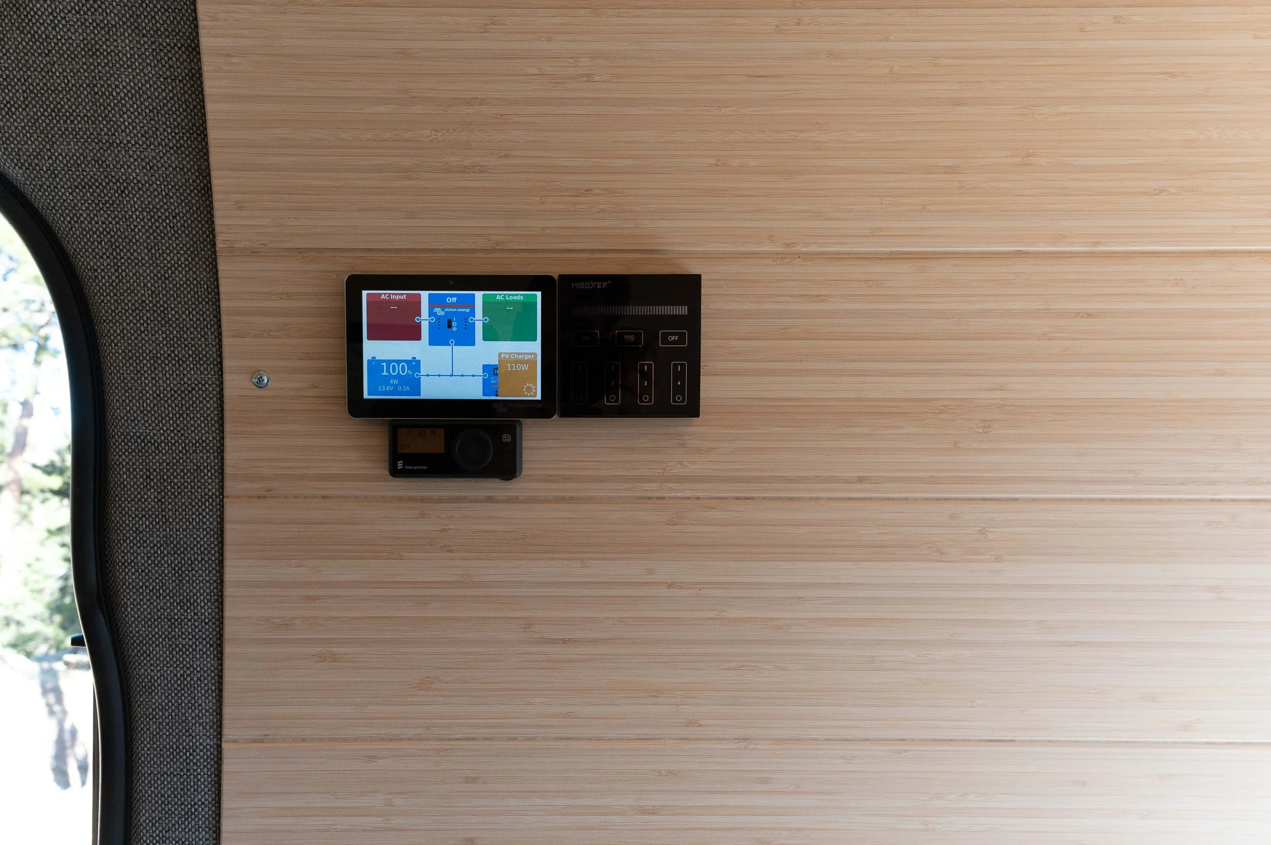 Wall with a small digital control panel, a black circuit breaker panel, and a small black device, all mounted on a wooden wall in a minimalist interior.