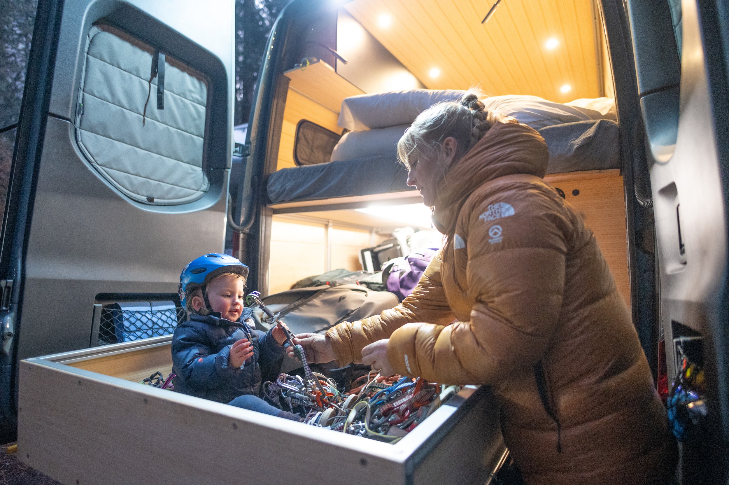 A woman helping a young child put on a climbing harness inside a camper van with a bed and wooden interior.