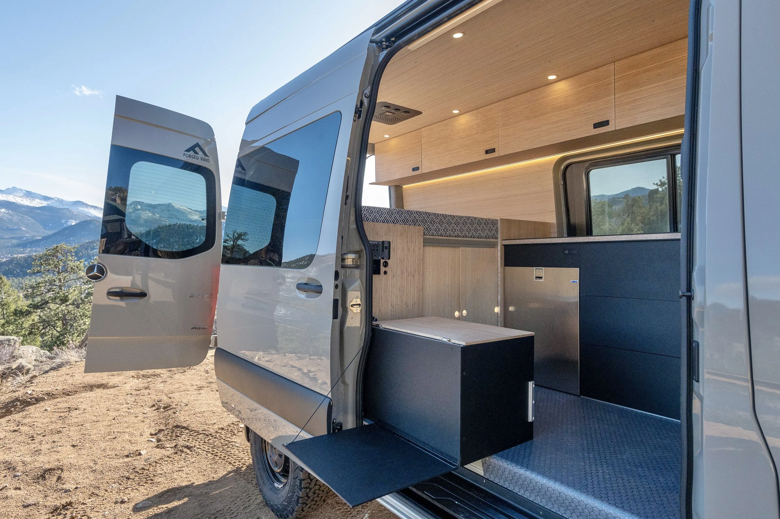 Interior of a camper van with wooden cabinets, black and gray storage units, and a window showing mountain scenery outside.