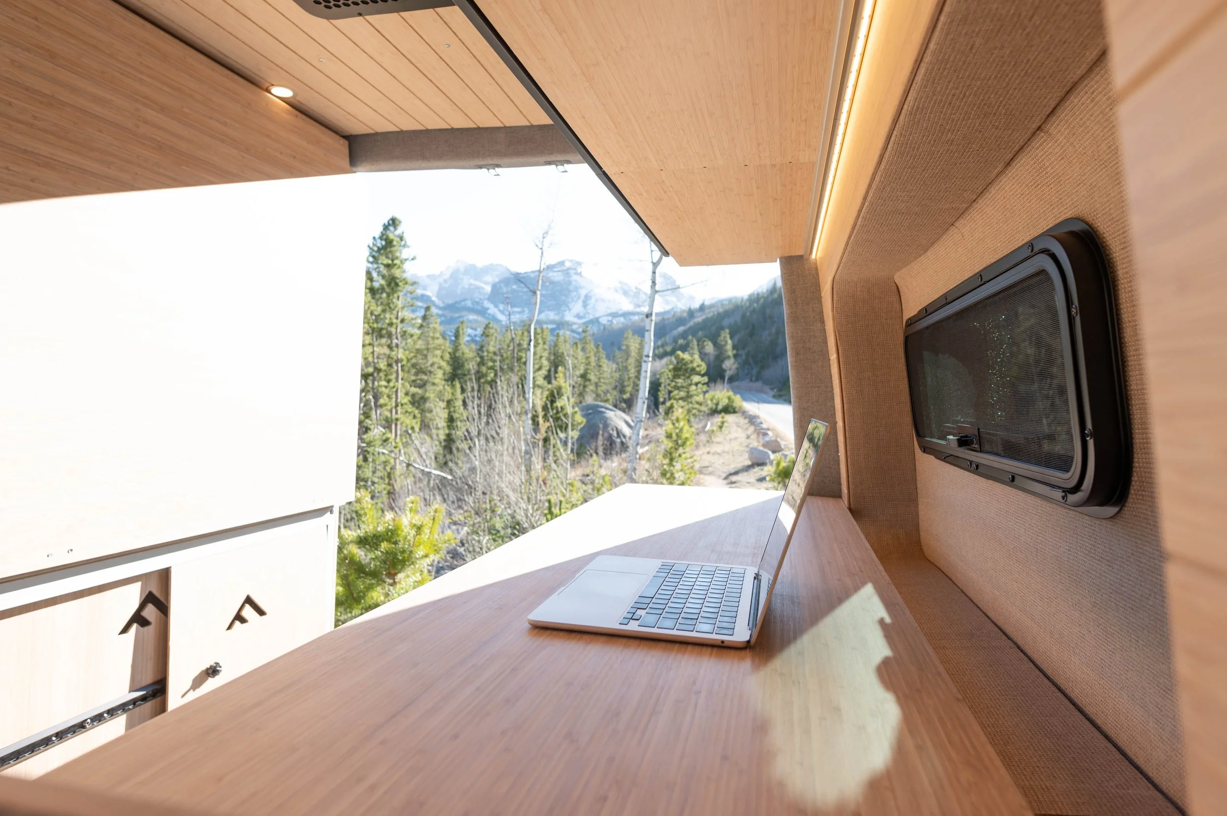 A minimalist wooden workspace with a laptop on a sloped wooden desk, overlooking a forest and mountain landscape through a window, with a small storage unit below.