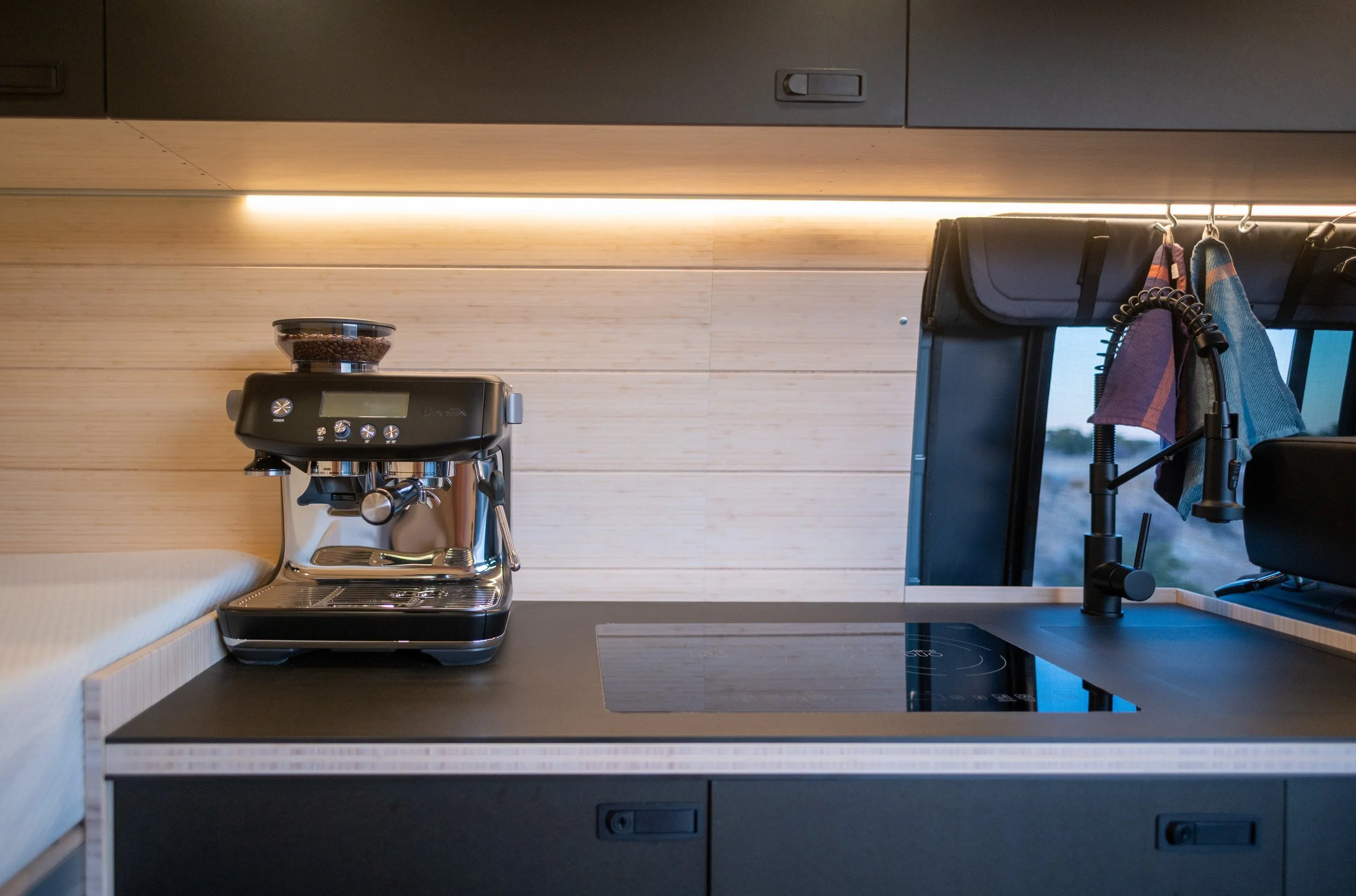 A modern kitchen countertop with a black espresso machine, a glass bowl of coffee beans on top, a sleek black sink with a tall black faucet, and towels hanging on hooks near a window with a view outside.
