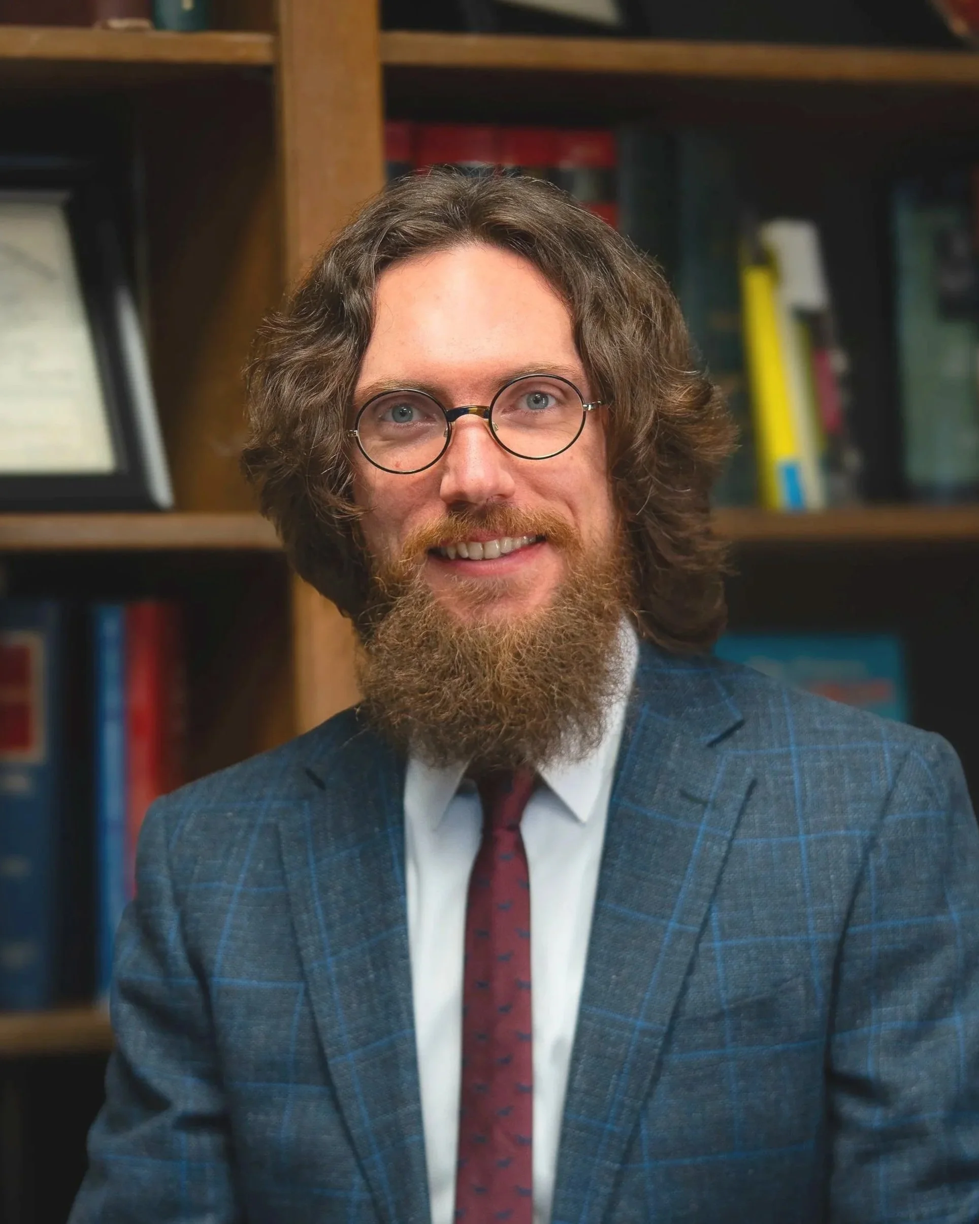 OMMA and OBNDD licensing Director Mason Pain, shown with long curly hair, glasses, and a beard, wearing a checkered blazer over a dress shirt with a subtle pattern.