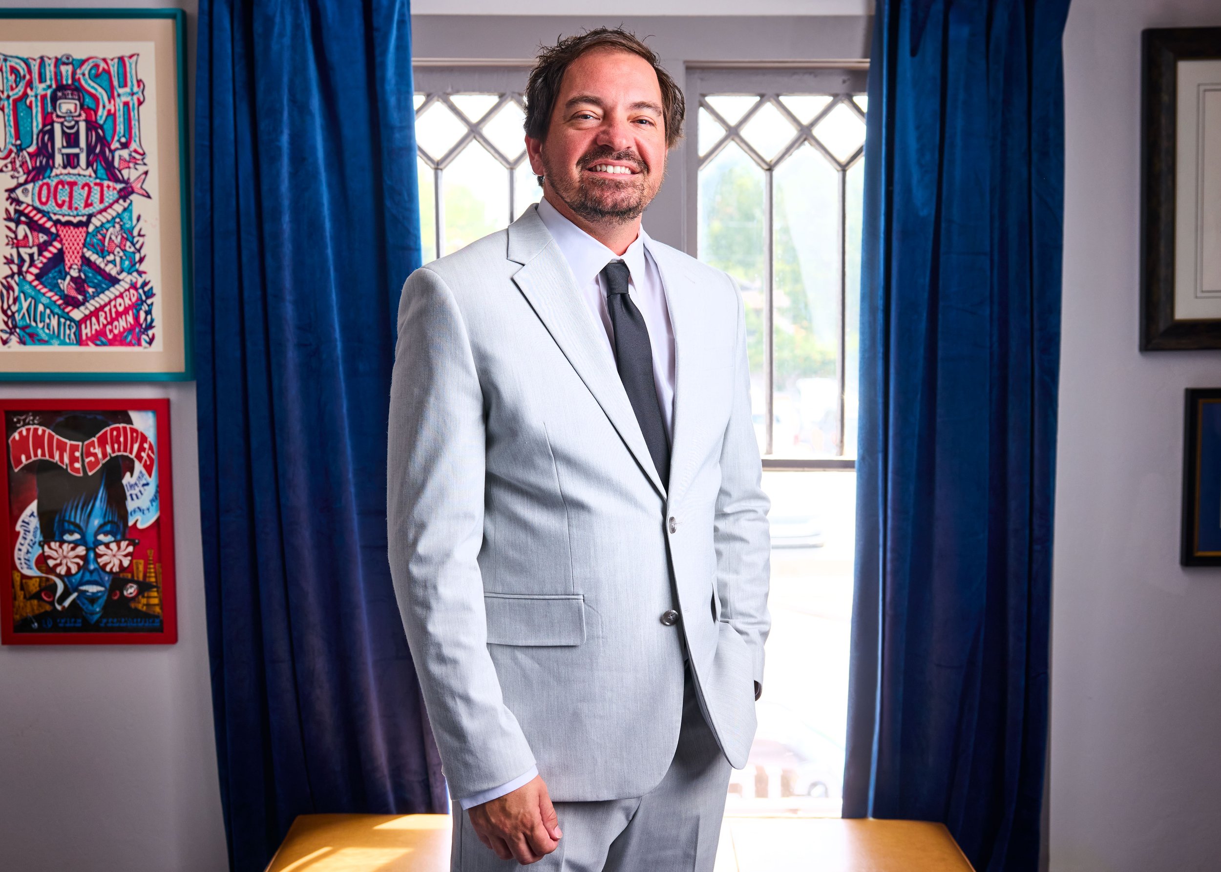 Oklahoma cannabis and criminal attorney Donald Gies sitting on a green leather tufted sofa, smiling, wearing a gray suit, blue shirt, and blue tie.