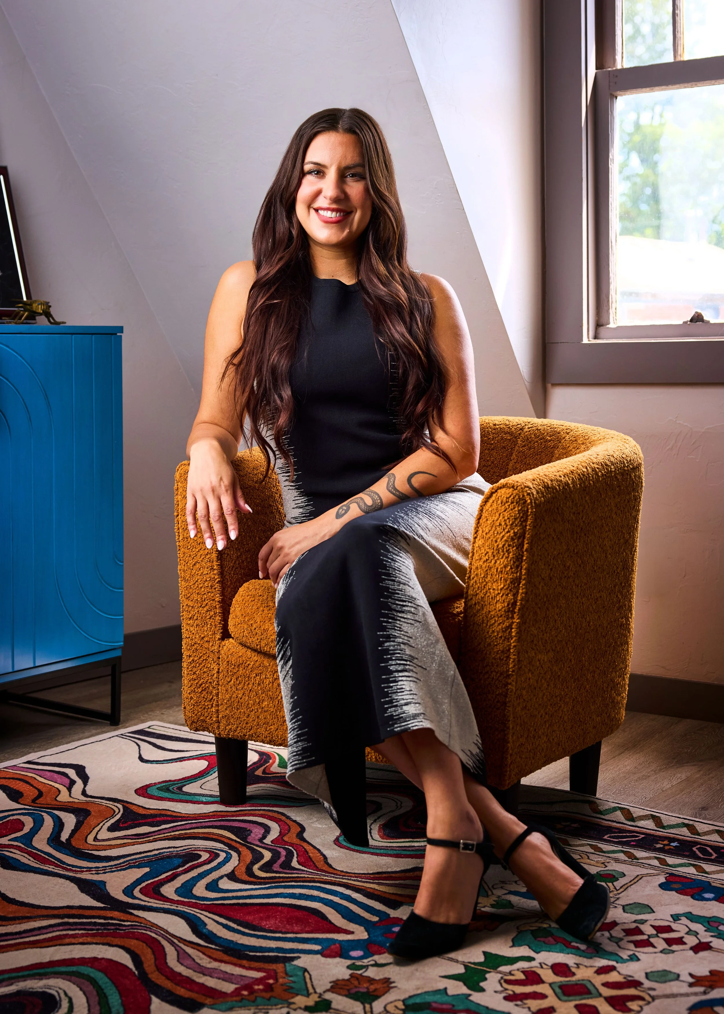 A woman with long dark hair and fair skin sitting in a black leather armchair in a room with soft lighting, a window with decorative grid pattern, and a lamp with a green shade in the background.