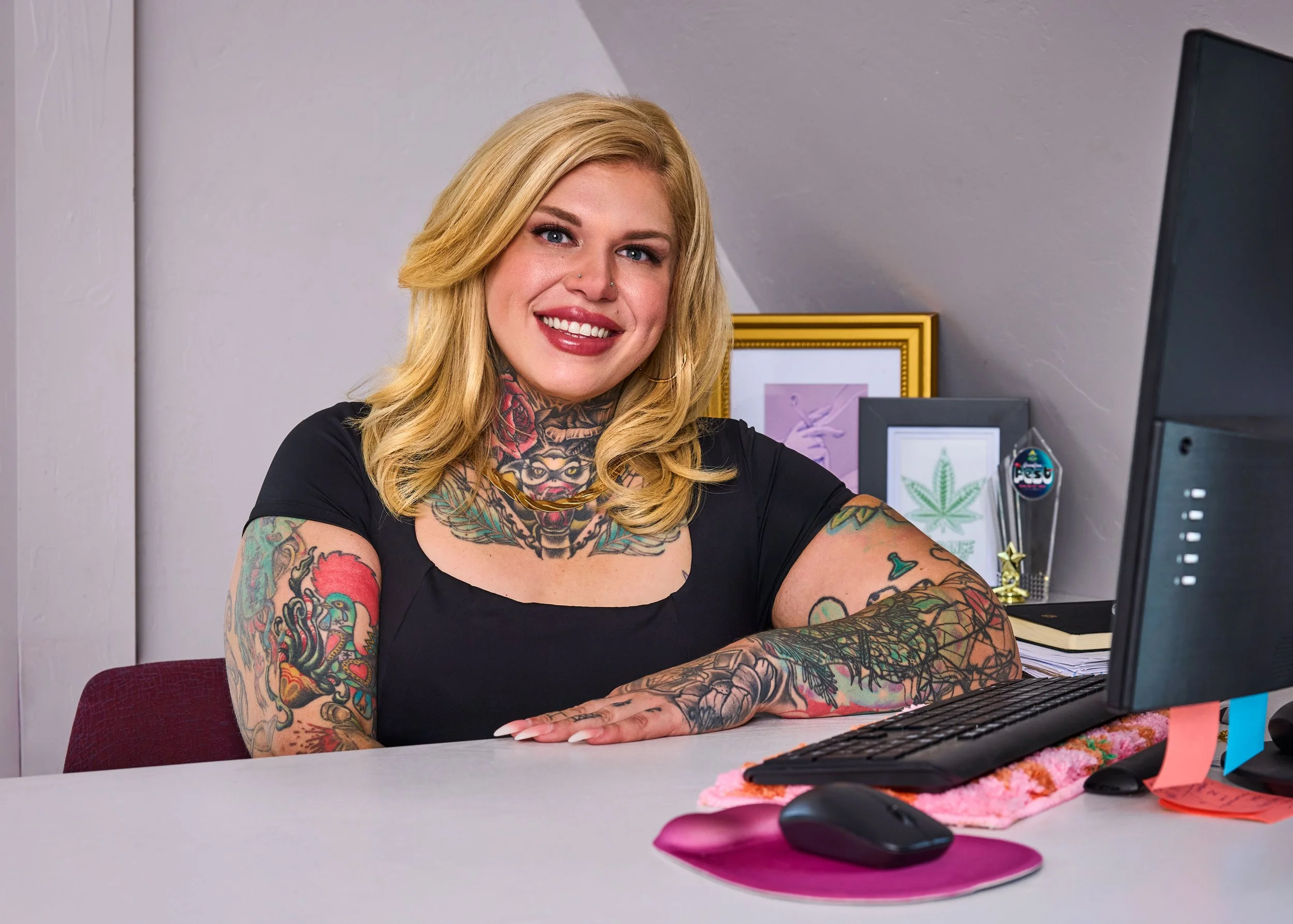 Close-up of a woman with long blonde hair, smiling, wearing a plaid blazer, a black top, and gold jewelry, with visible tattoos on her chest.