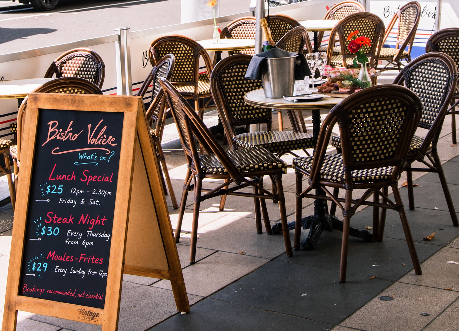 Outdoor patio of a restaurant with chairs and tables, a blackboard menu stand, and a large striped umbrella with the writing 'Bistro Valée' on it. Cars are visible on the street and trees in the background.