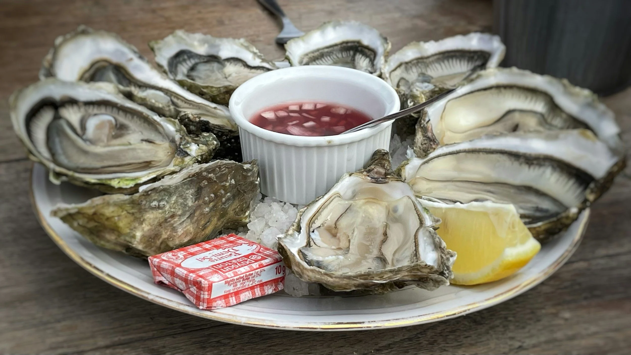A plate of raw oysters on ice with a lemon wedge and a small cup of cocktail sauce, labeled 'Bistro Voilier'.