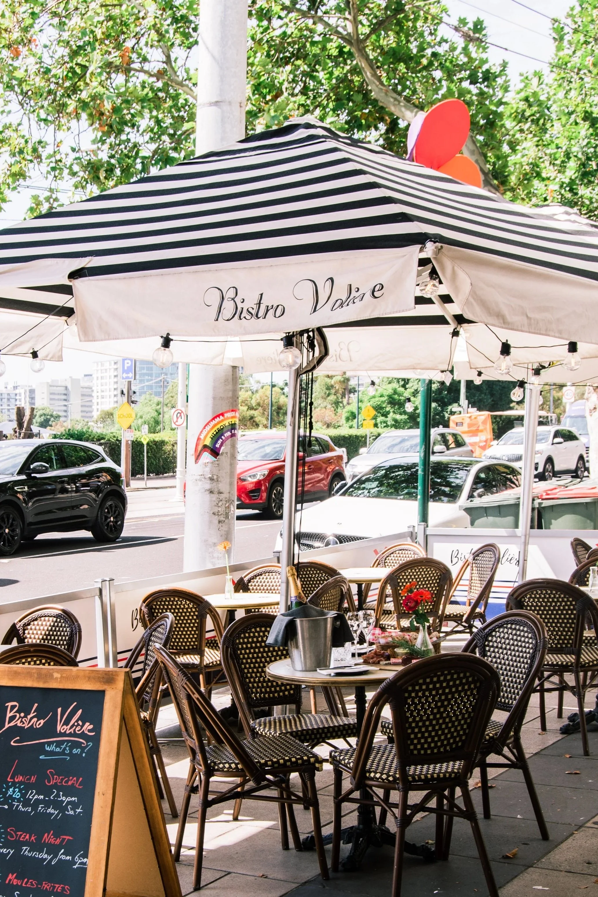 Outdoor patio of a restaurant with chairs and tables, a blackboard menu stand, and a large striped umbrella with the writing 'Bistro Valée' on it. Cars are visible on the street and trees in the background.