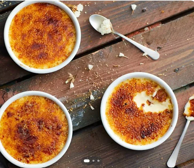 Three bowls of crème brûlée with caramelized sugar on top, two spoons with some leftover custard, and scattered sugar and crumbs on a wooden table.