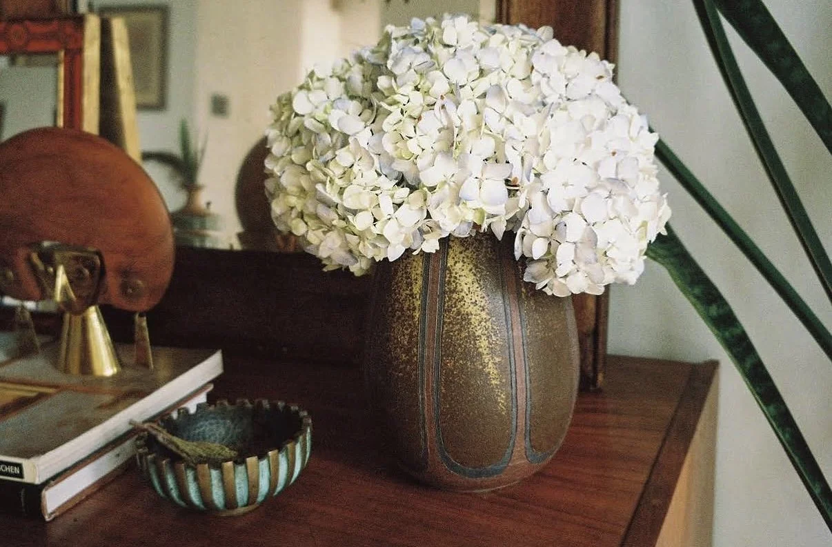 A large ceramic vase with a metallic finish and decorative lines, holding a bouquet of white hydrangea flowers on a wooden table.