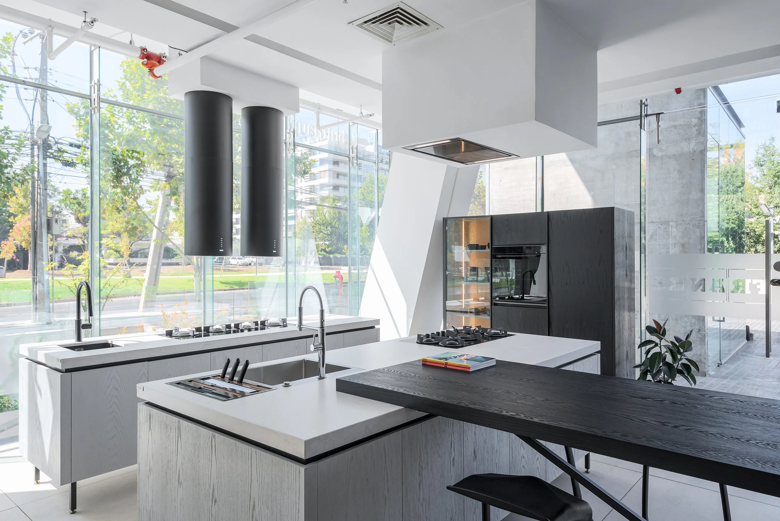 Modern kitchen with white island counters, black cabinets, high ceilings, large floor-to-ceiling windows, and outdoor trees visible outside.