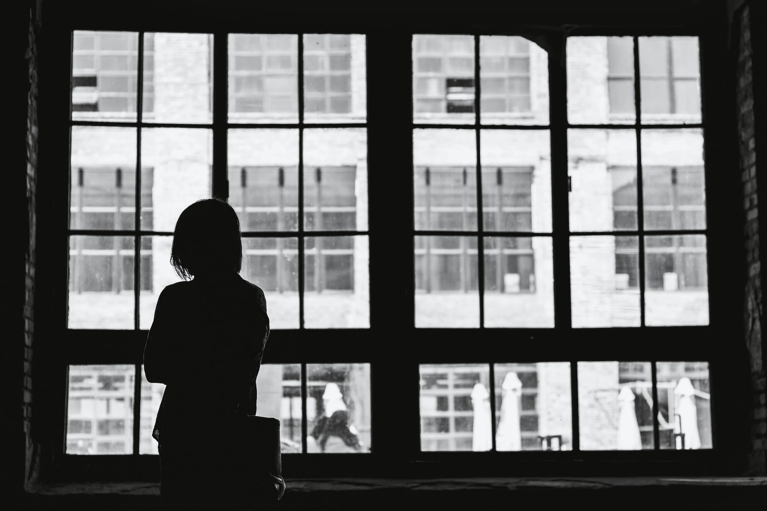 Silhouette of a woman standing in front of a large industrial-style window, with an urban building visible outside.