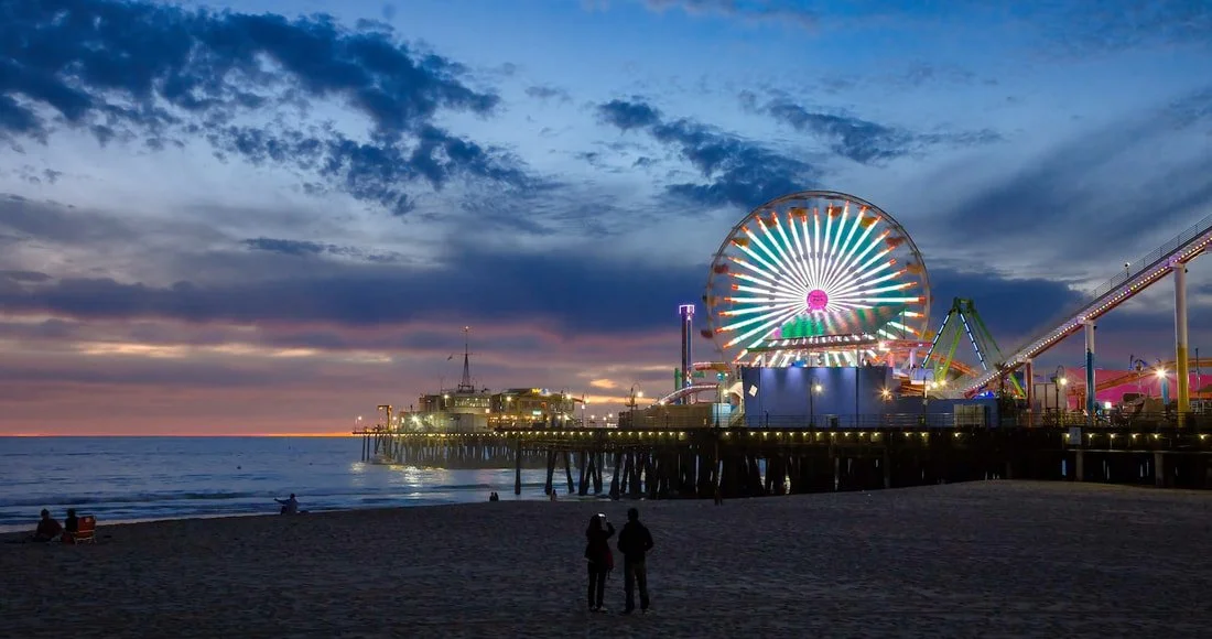 A nighttime view of a pier with a brightly lit Ferris wheel, roller coaster, and other amusement rides, with a cloudy sky and a few people on the beach in the foreground.
