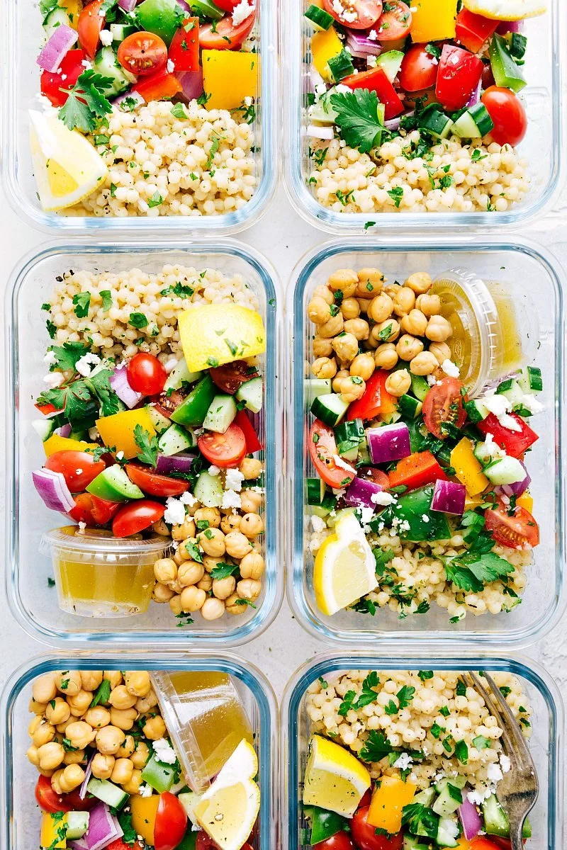 Glass meal prep containers filled with healthy Mediterranean salad ingredients including cherry tomatoes, cucumber, bell peppers, red onion, chickpeas, couscous, lemon wedges, and parsley, with small containers of dressing.