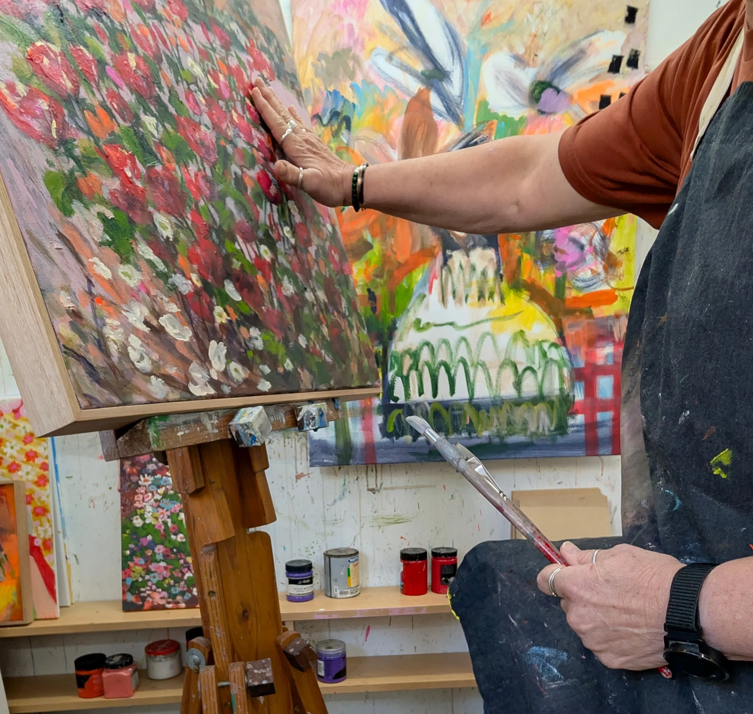 This is a close-up of artist Claire Cummack working at an easel in her art studio. One hands is on the painting, the other holding a brush. Art supplies such as jars of paint are visible on the shelves in the background.